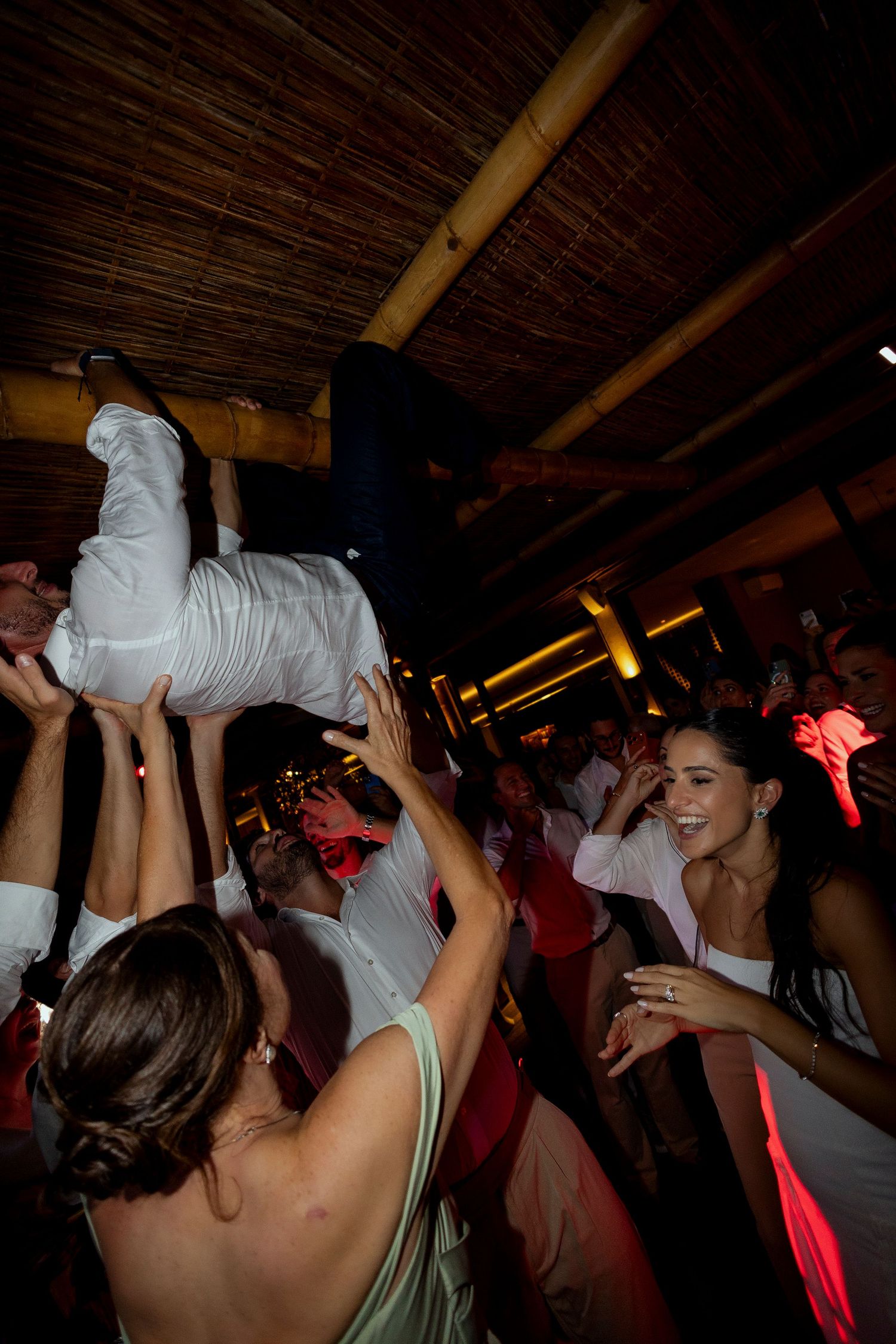 Crowd lifting a man in white pants overhead at a lively wedding reception dance floor with red lighting.