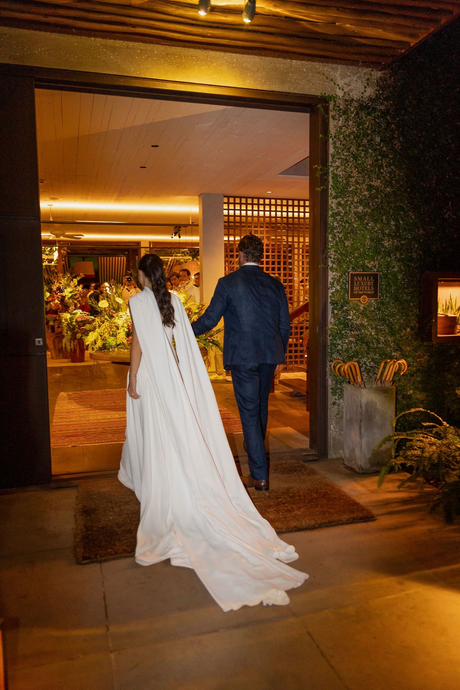 A bride in a white gown with long train and groom in navy suit hold hands entering a warmly lit venue.