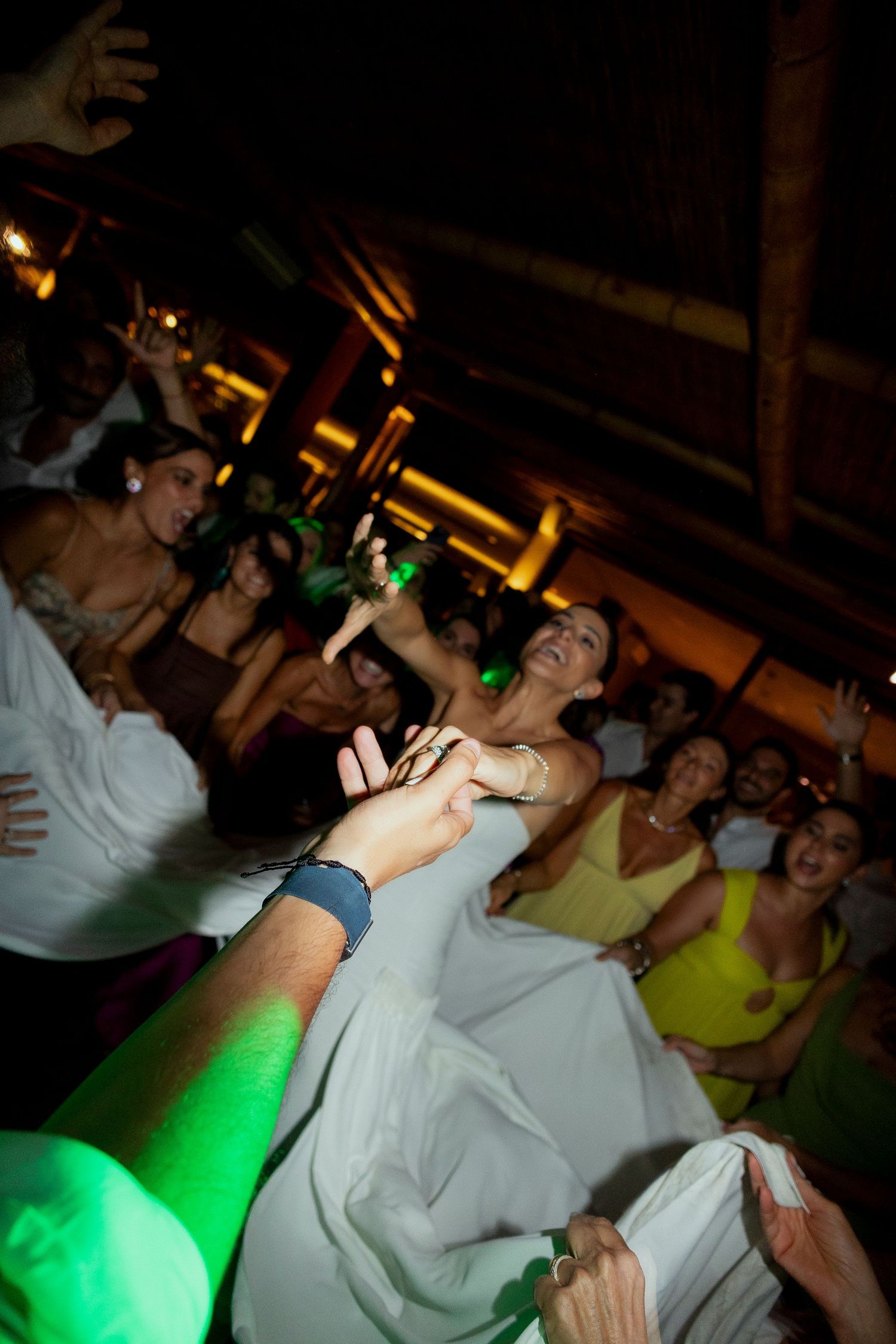 Bride laughing joyfully while being lifted by guests during a lively wedding reception celebration indoors.