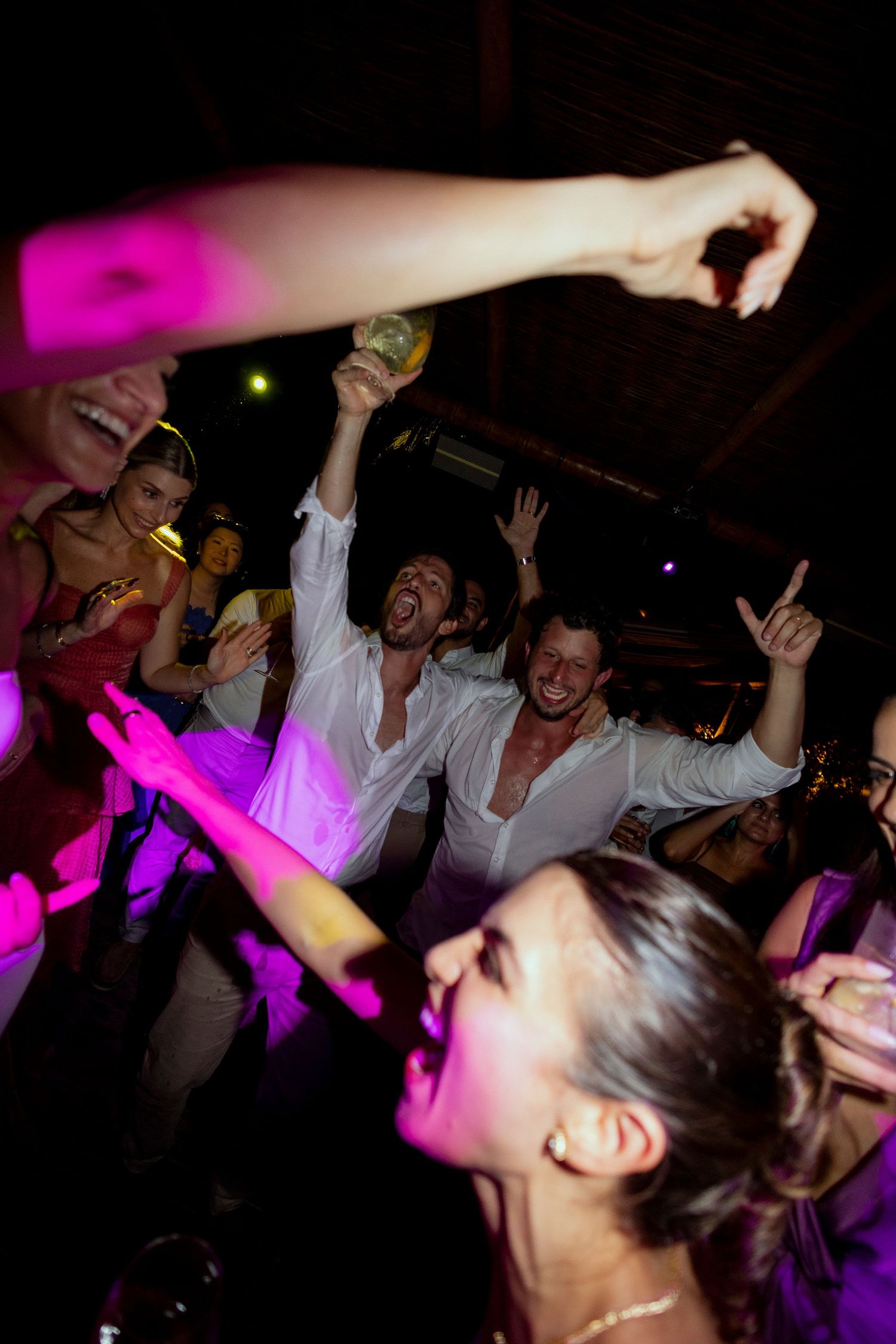Energetic party crowd dancing and celebrating under colorful pink and purple lights at a lively nighttime event.
