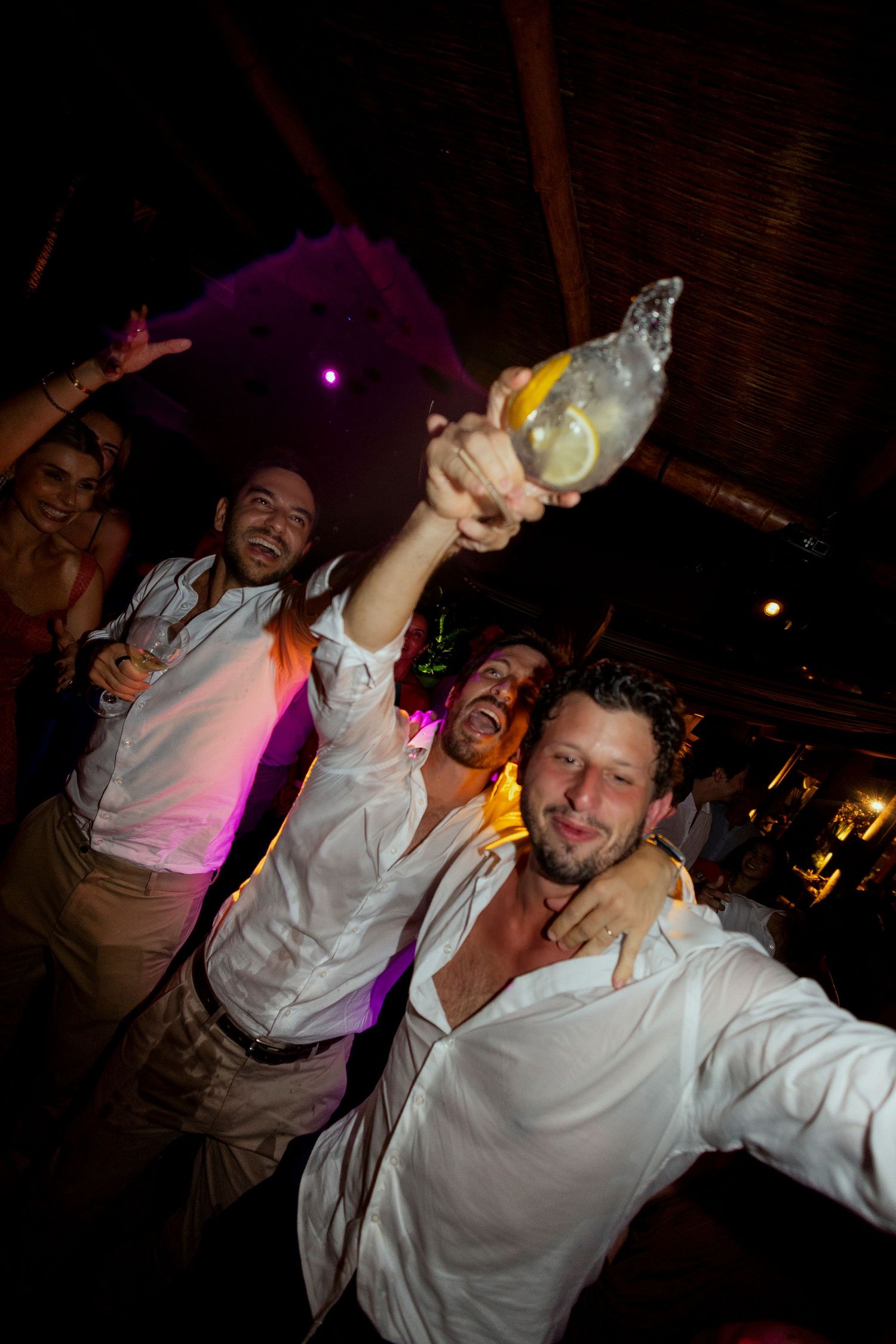 Group of men in white shirts celebrating at a nightclub, raising drinks and taking a selfie under purple lights.