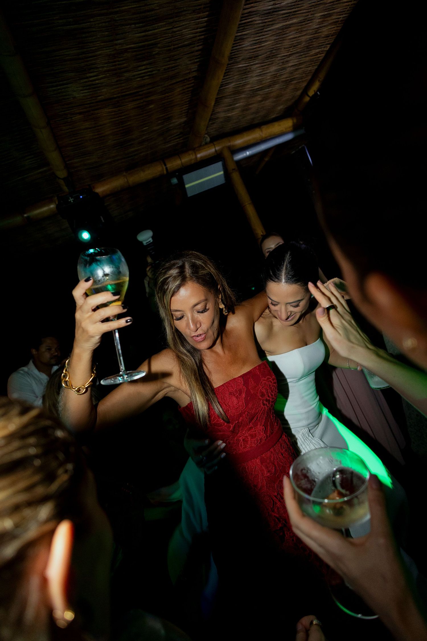 Women in red and white dresses dancing at a party, holding cocktail glasses under green lights.