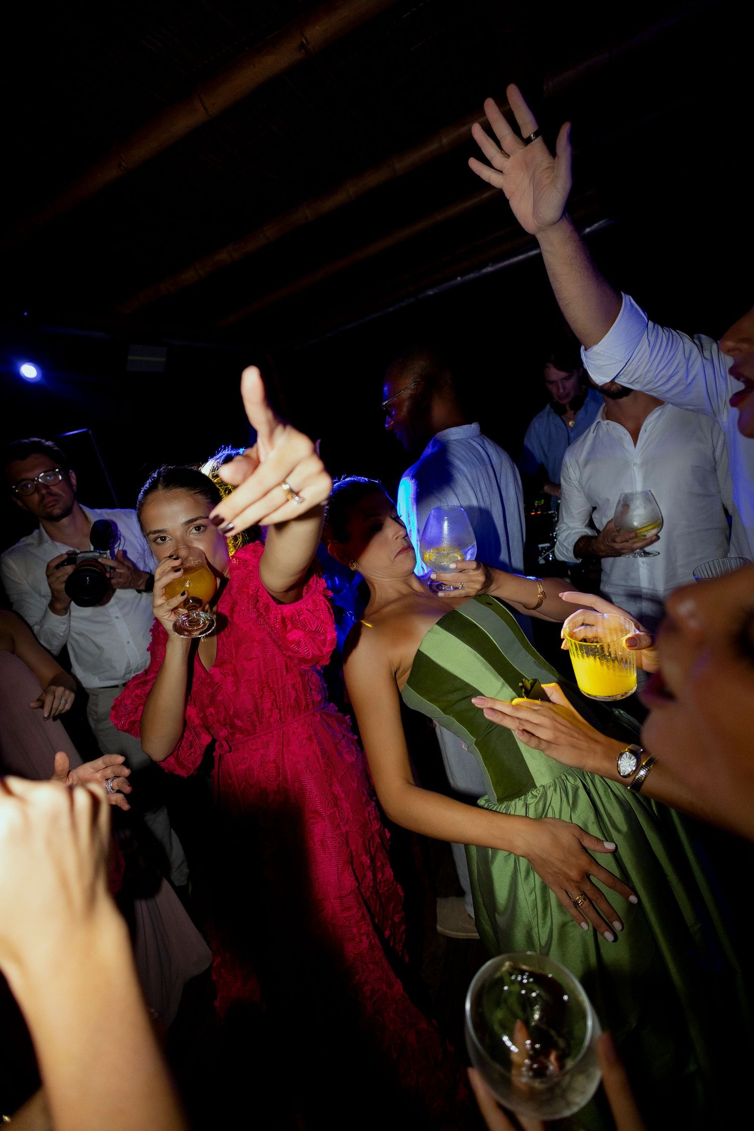 People dancing and celebrating at a party, holding drinks with hands raised in a dark venue.