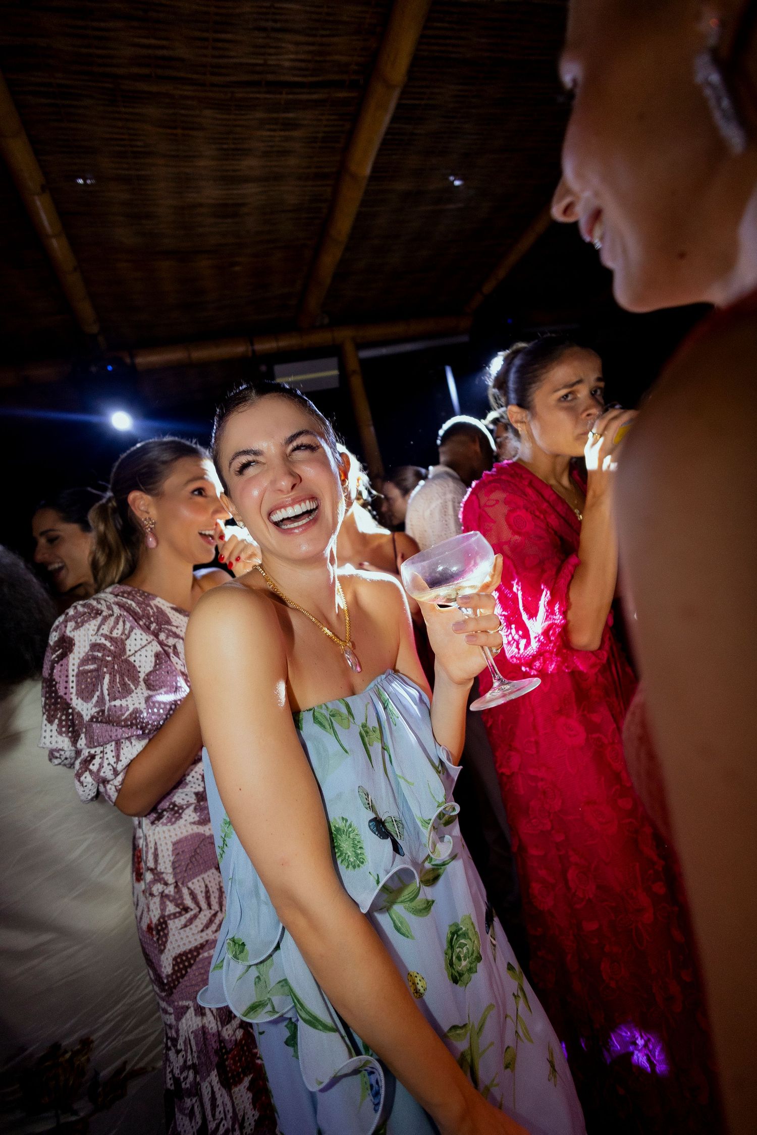 Women laughing and socializing at an outdoor evening party, holding wine glasses while wearing colorful dresses.