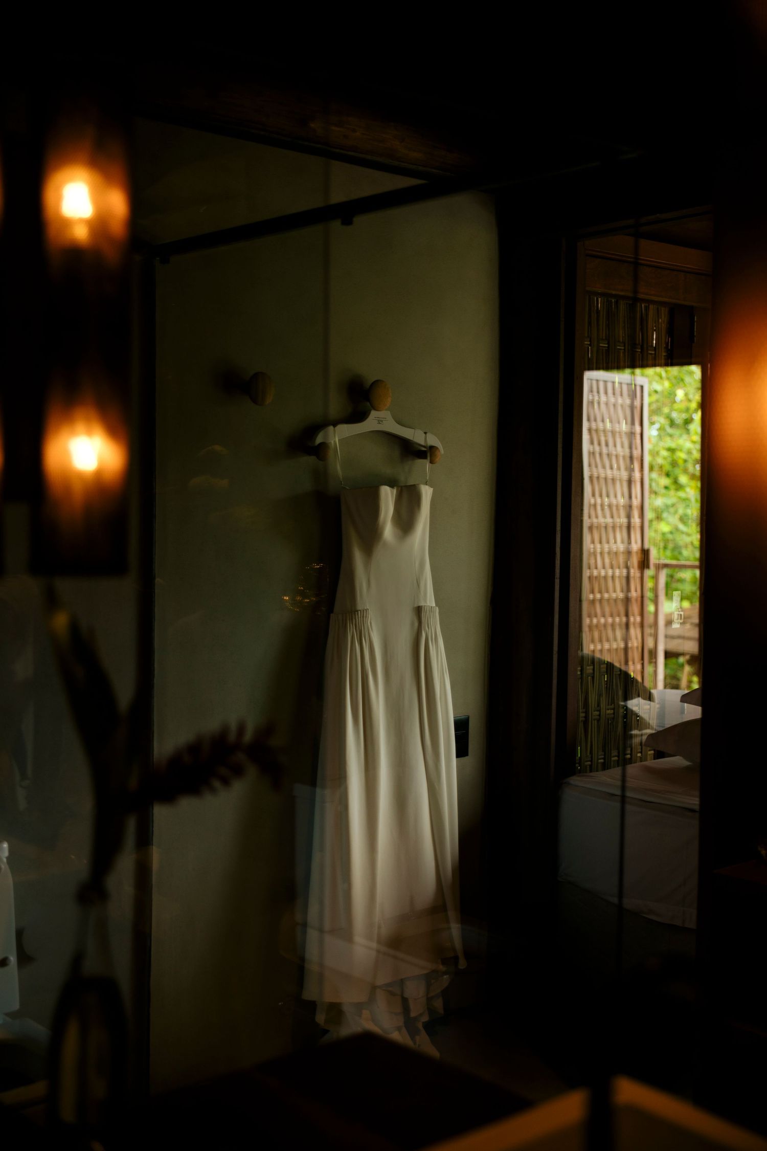 White wedding dress hanging on a door in a dimly lit room with warm glowing lights and greenery visible outside.