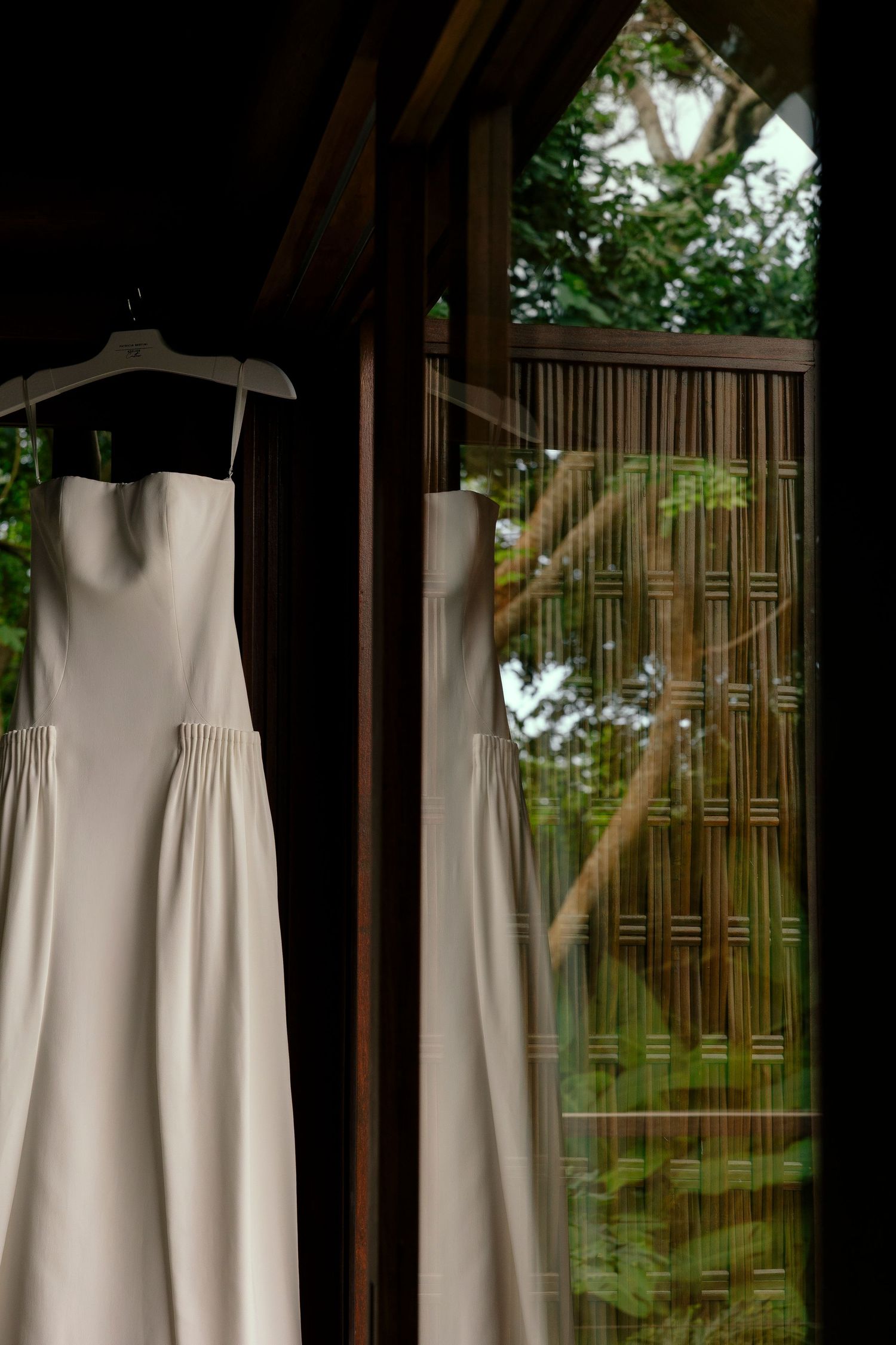 Elegant white strapless wedding dress hanging on a hanger near a wooden window with lush green garden view.