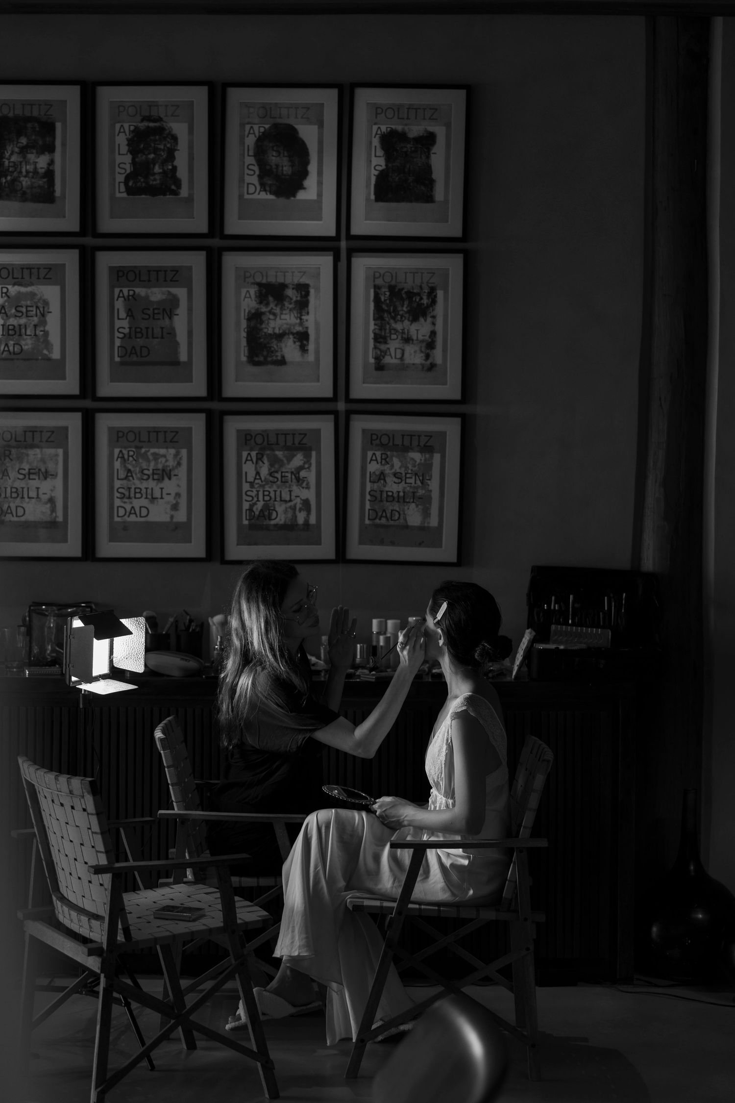 Black and white photo of a makeup artist applying makeup to a bride in a dimly lit room with framed art on the wall.