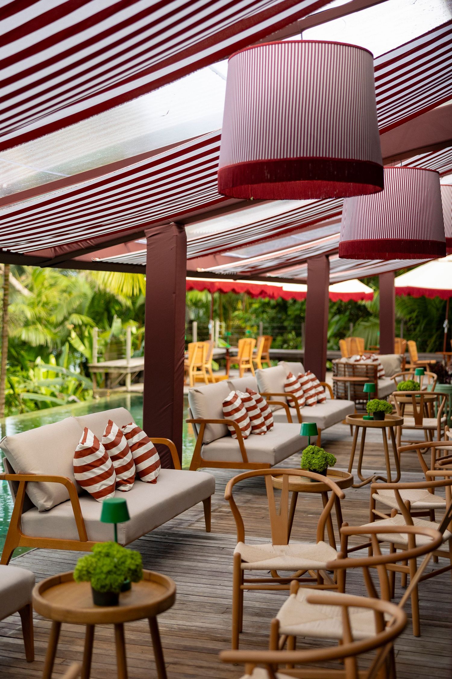 Elegant outdoor lounge with red striped canopy, pendant lamps, wooden furniture, striped cushions, and lush tropical greenery.