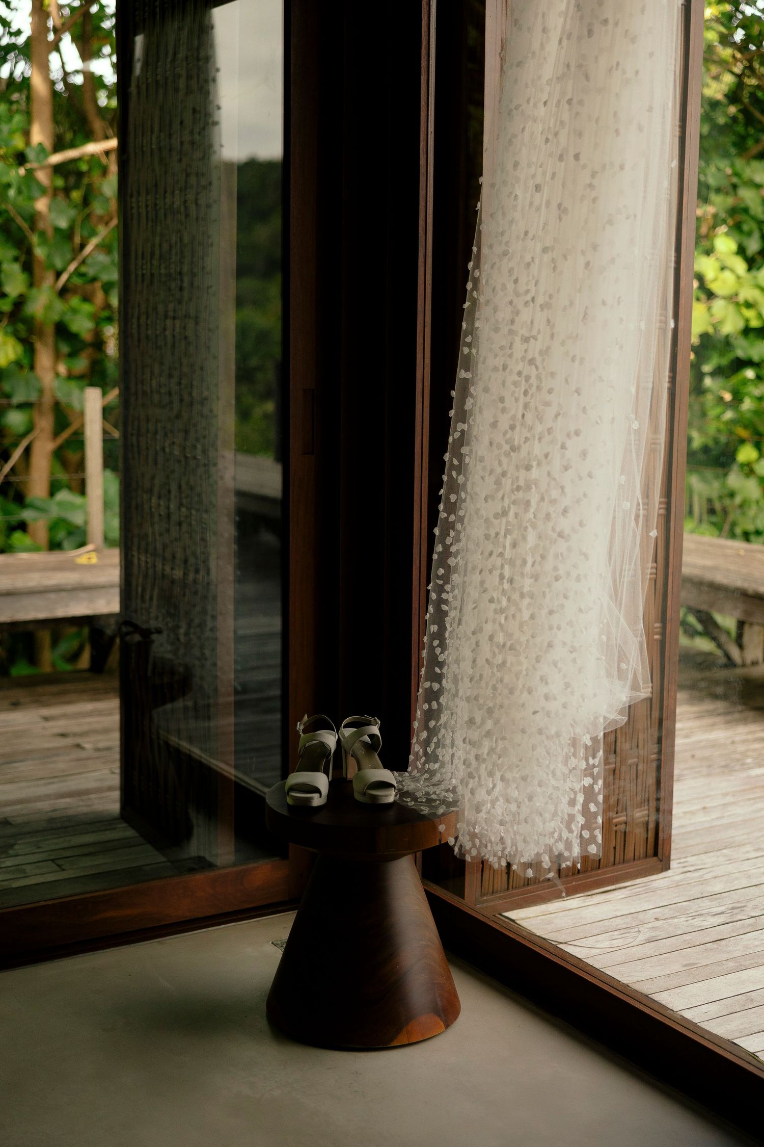 Wedding dress and heels displayed on a wooden stand by glass doors with sheer curtains and lush greenery outside.