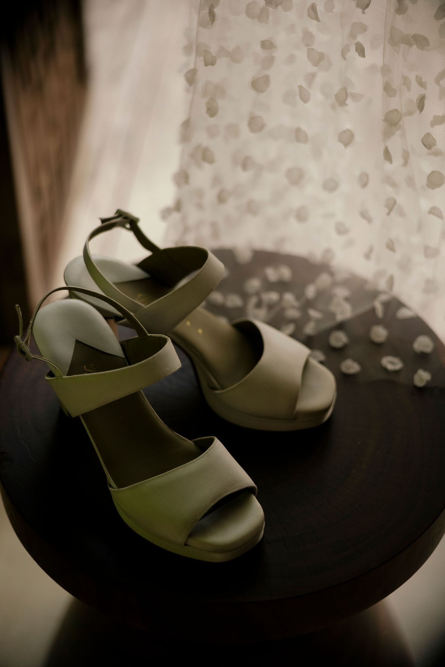 Elegant olive green platform wedge sandals displayed on a dark wooden table with sheer floral curtain backdrop.