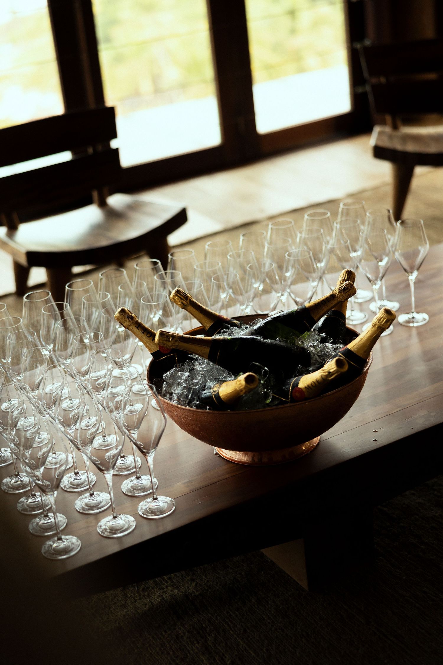Champagne bottles chilling in ice bowl surrounded by elegant empty wine glasses on wooden table.