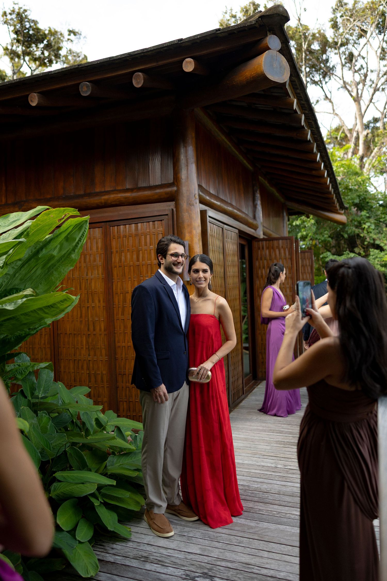 Couple posing for photo outside wooden tropical venue, woman in red gown and man in navy blazer, photographer capturing moment.