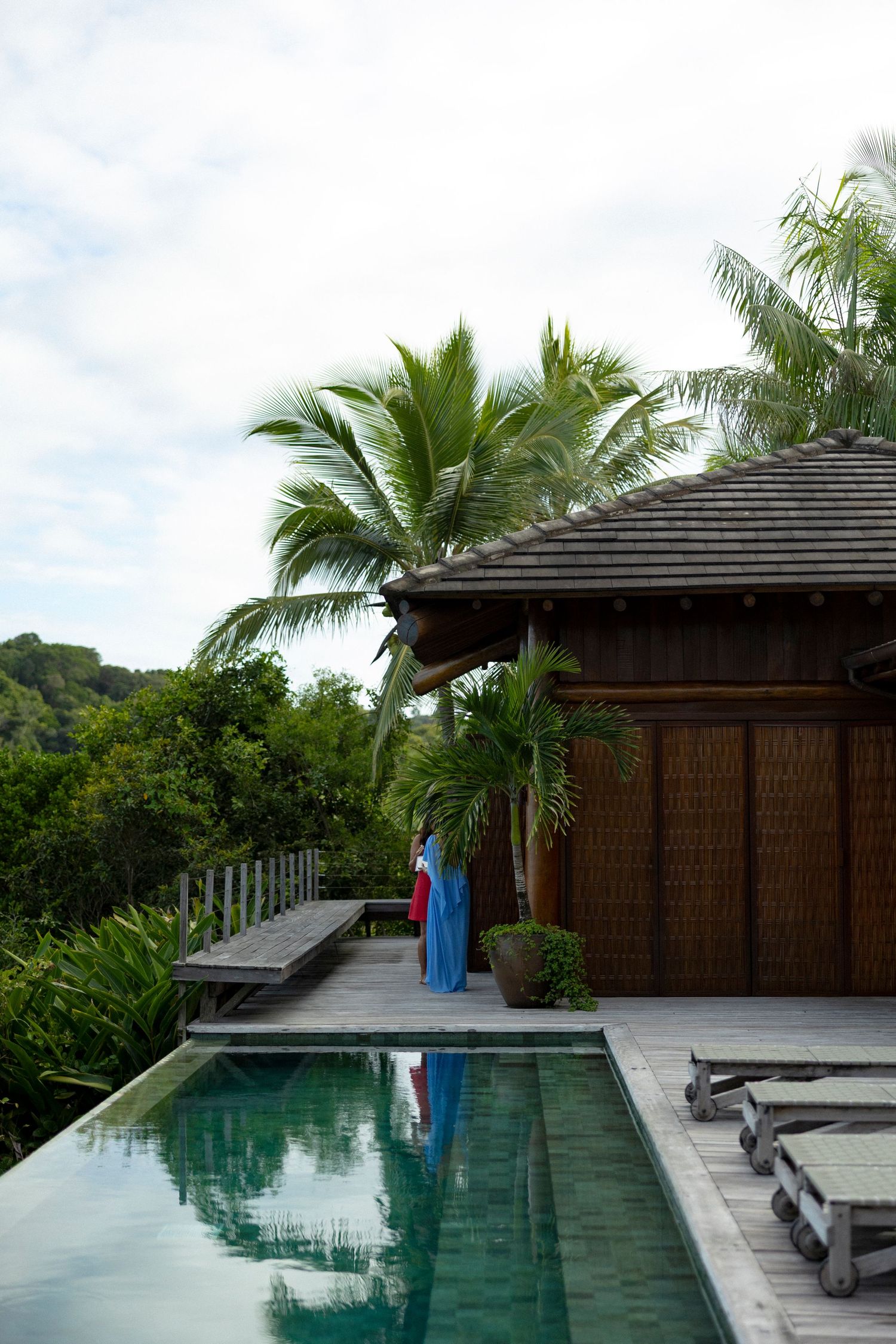 Luxury tropical villa with infinity pool, wooden bungalow, palm trees, and lush greenery under a cloudy sky.