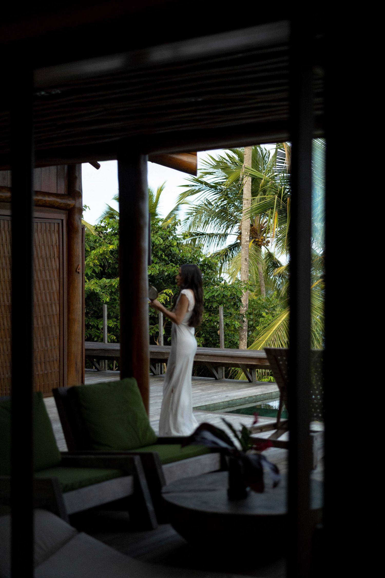 Woman in white dress standing on tropical villa terrace surrounded by lush palm trees and greenery.