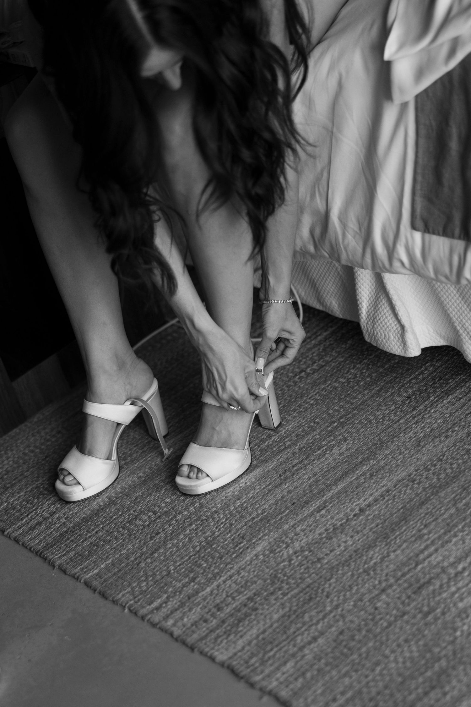 Bride being helped into white heeled sandals on a textured rug in a black and white wedding preparation photo.