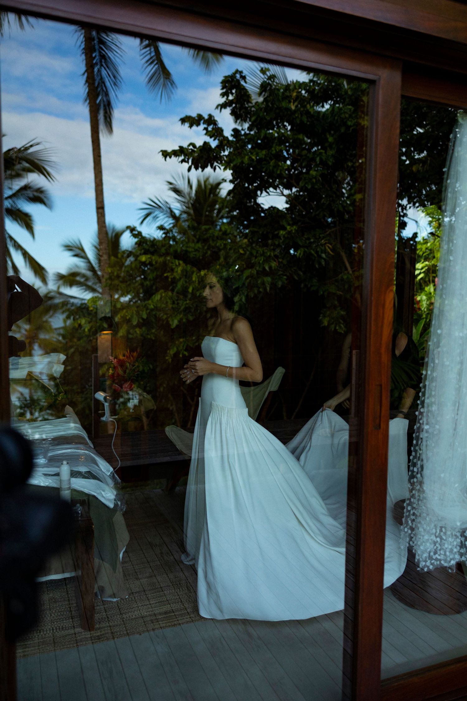 Elegant bride in a flowing white strapless gown viewed through a glass door with tropical palms.