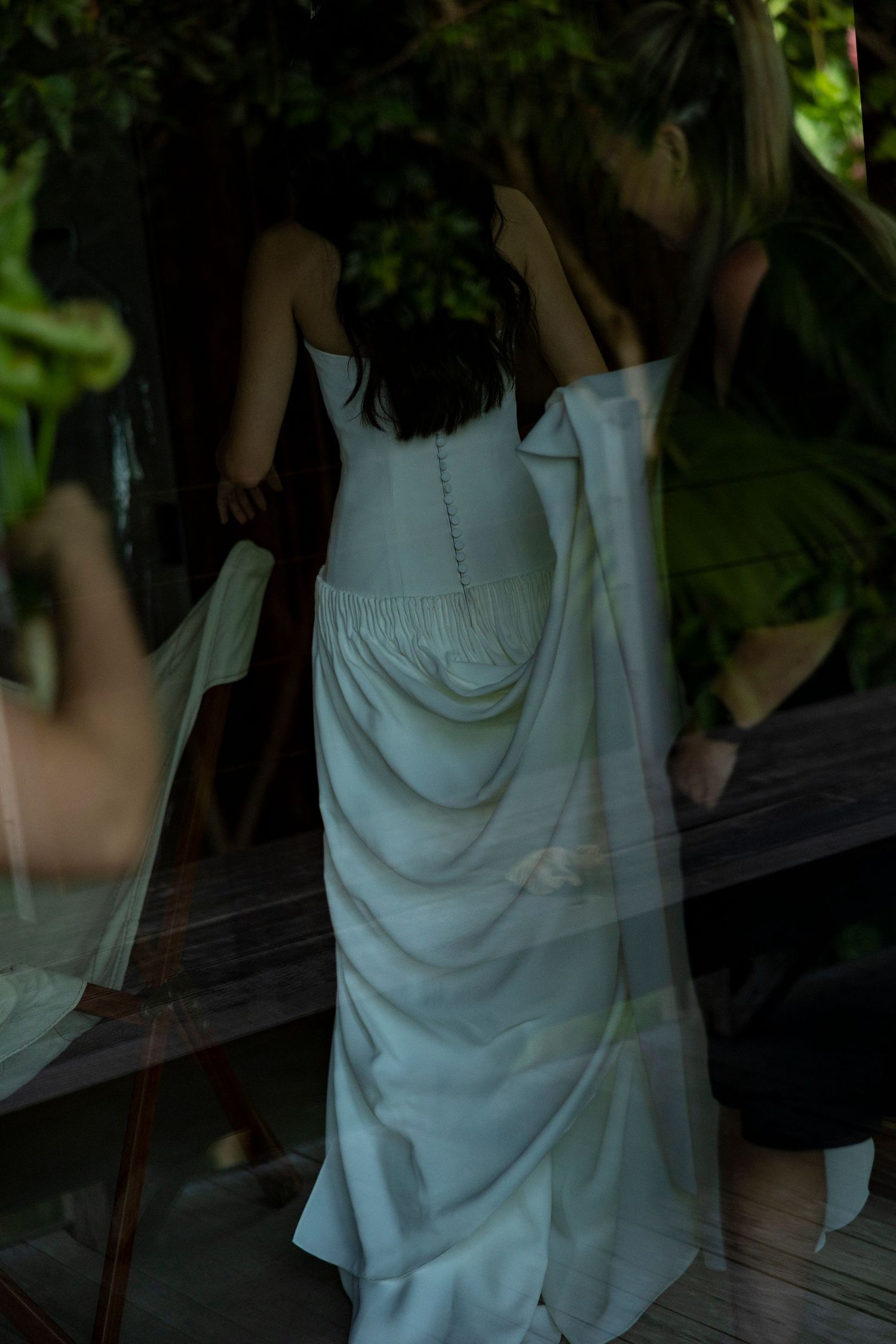 Bride in white wedding gown viewed from behind, standing near wooden table surrounded by lush tropical greenery.