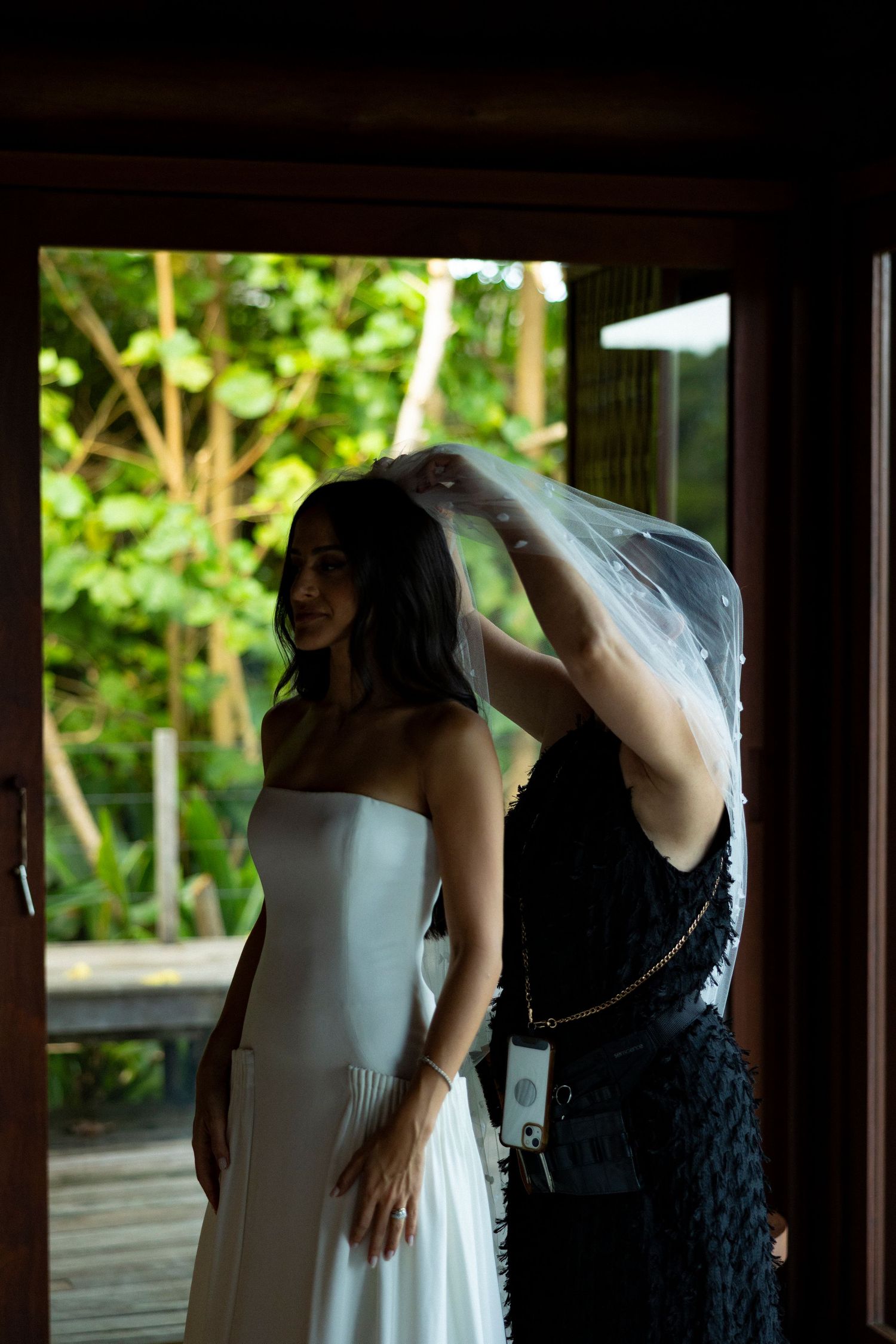 A bride in a white strapless gown smiles as her bridesmaid places a veil on her head near a sunlit doorway.