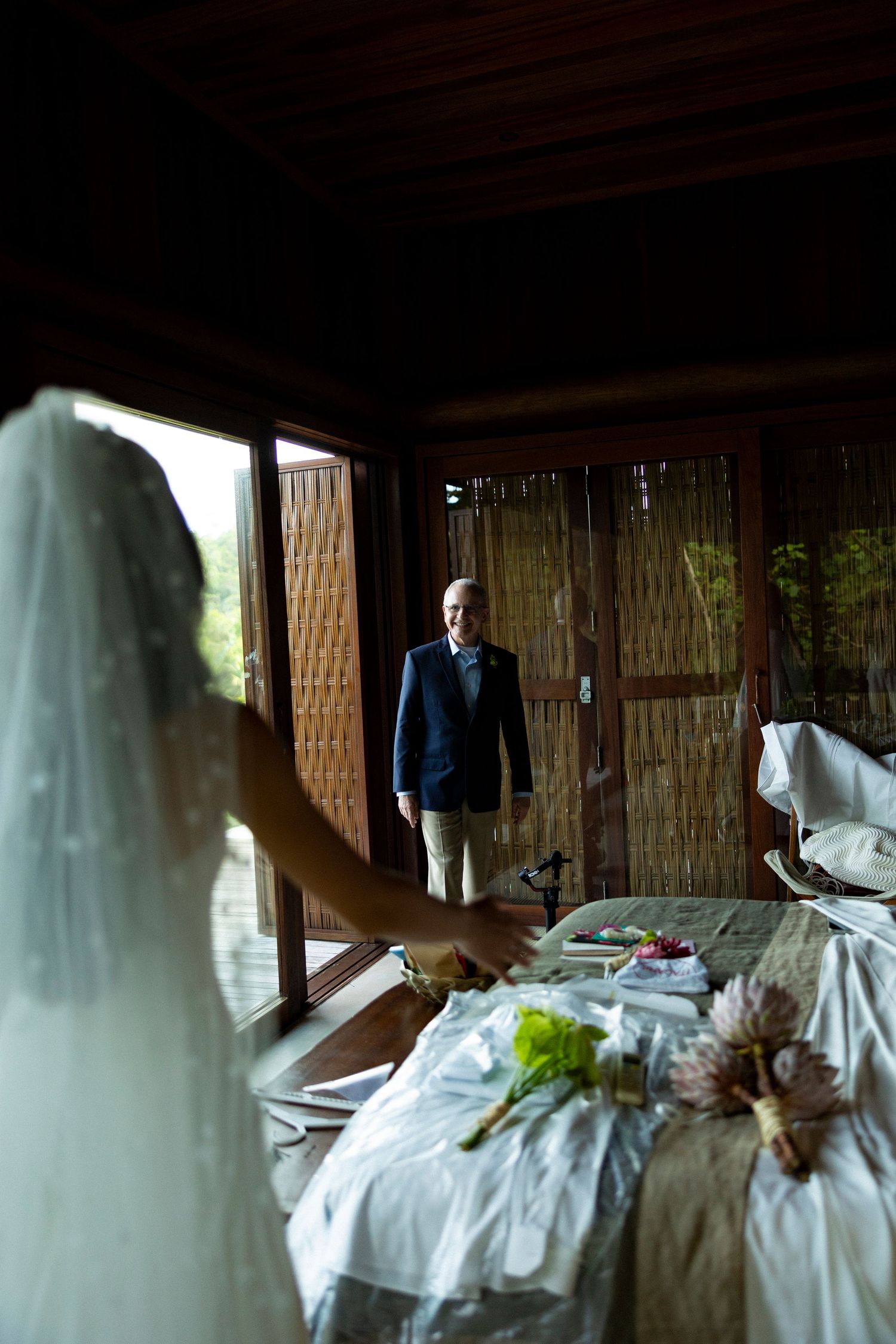 Bride in white veil faces her father in a blue suit during an emotional wedding day first look moment indoors.