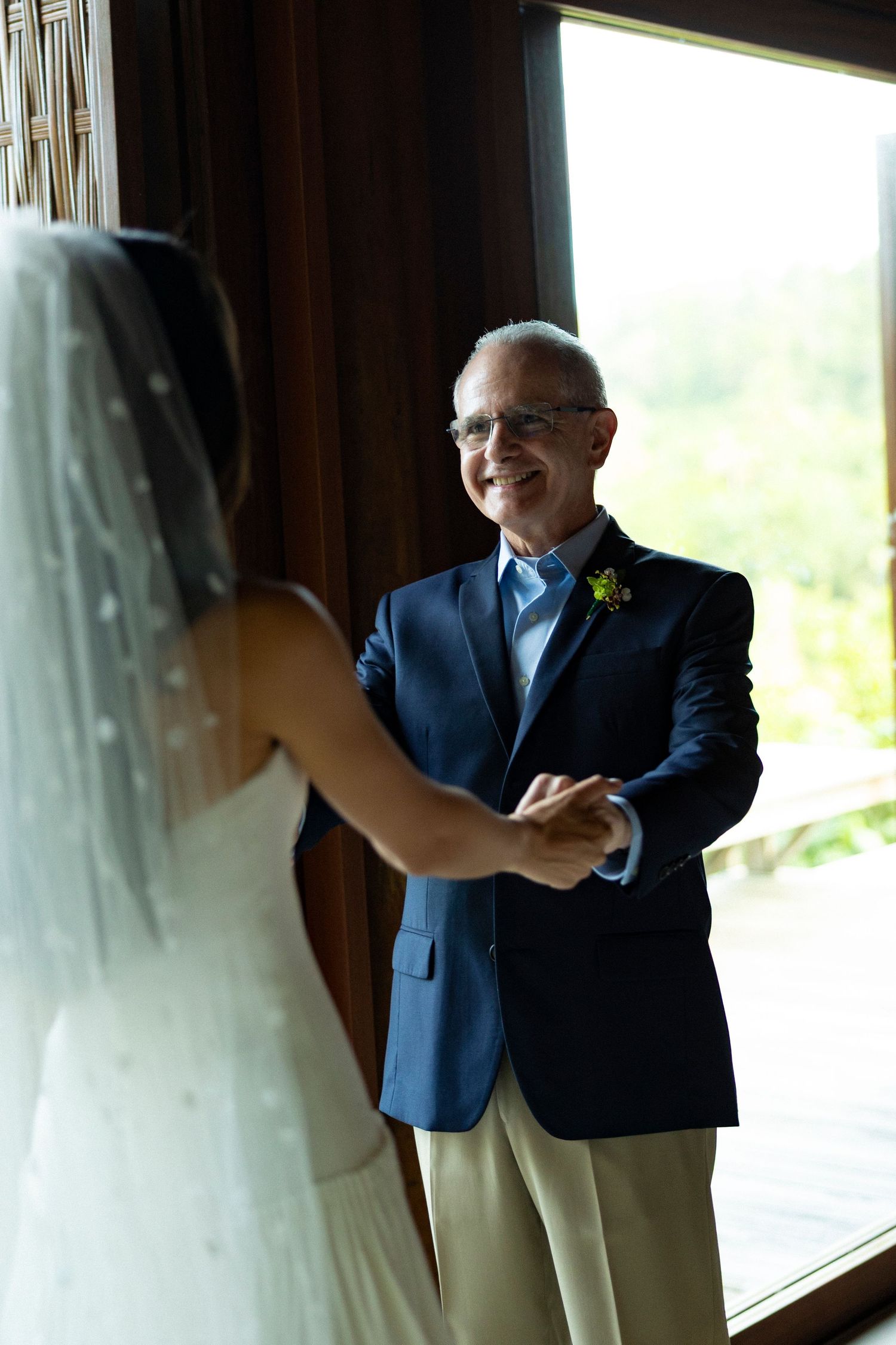 An older man in a navy suit smiles warmly while holding hands with a veiled bride during a first look moment.