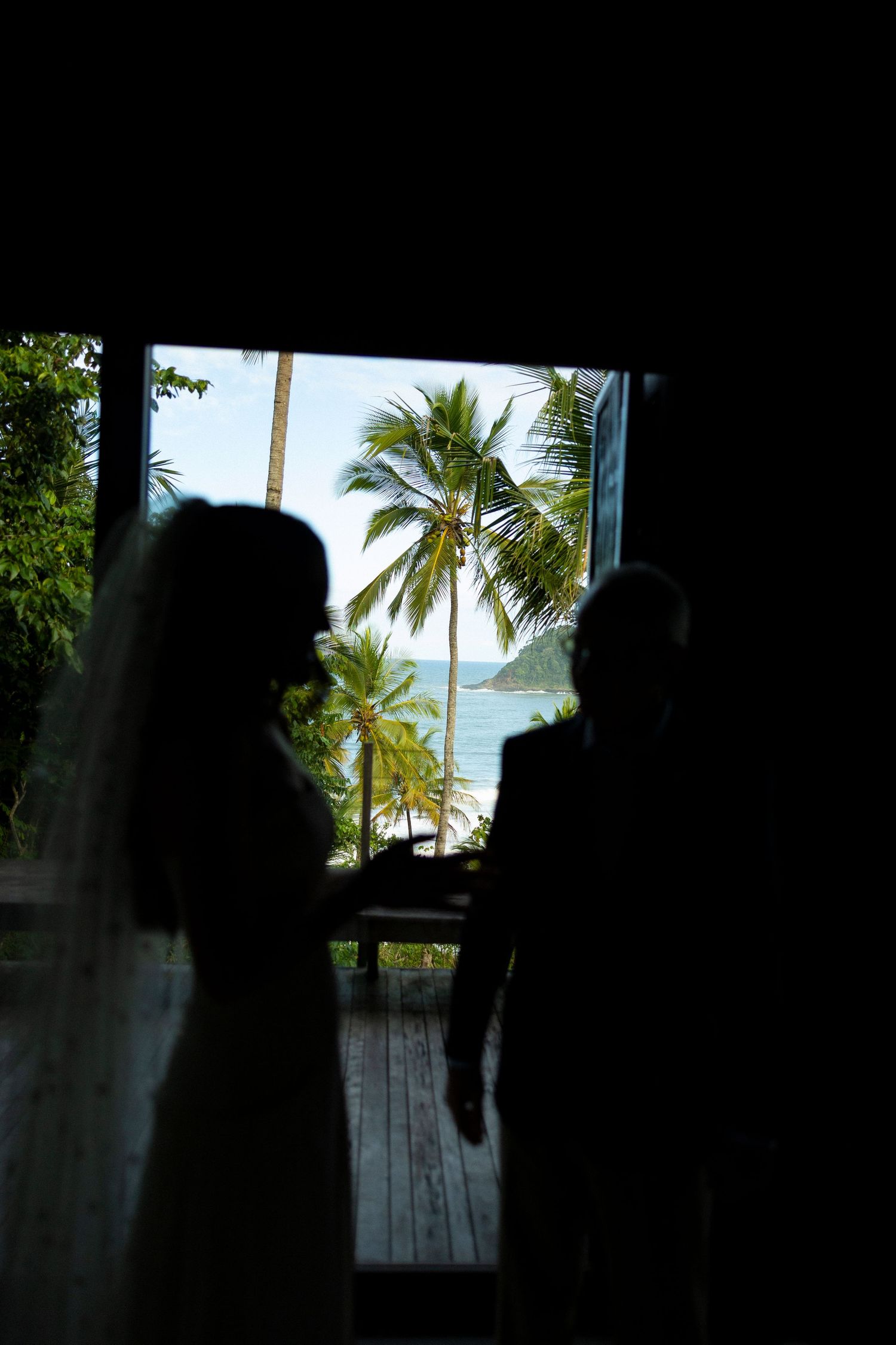 Silhouette of a bride and groom standing in a doorway with tropical palm trees and ocean view behind them.