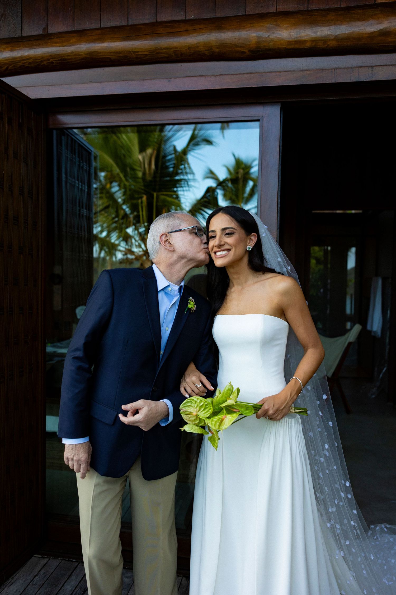 Older man in navy suit kisses smiling bride in strapless white gown holding green anthurium bouquet outdoors.