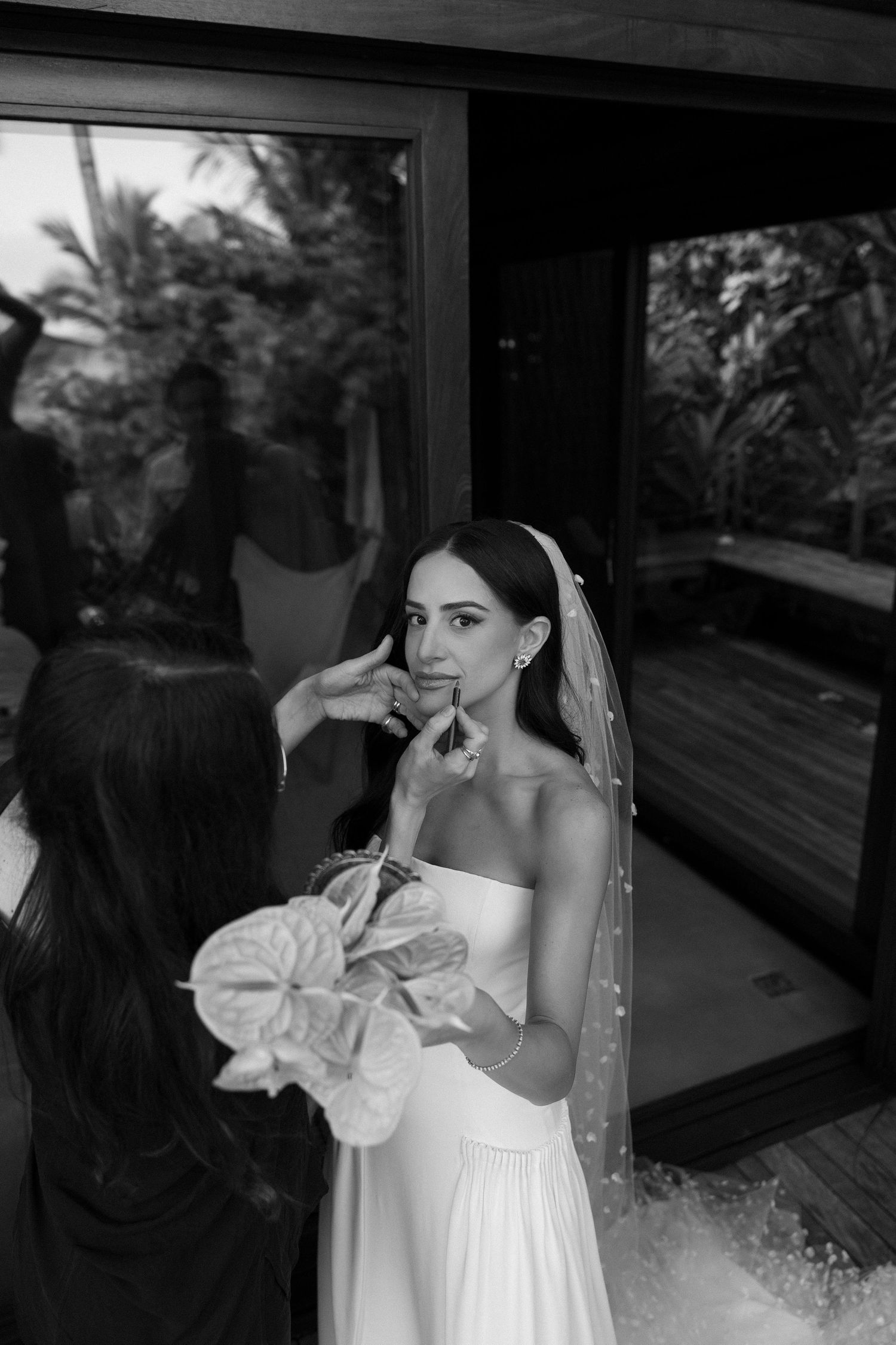 Black and white photo of a bride in a strapless gown and veil getting makeup applied before her wedding ceremony.