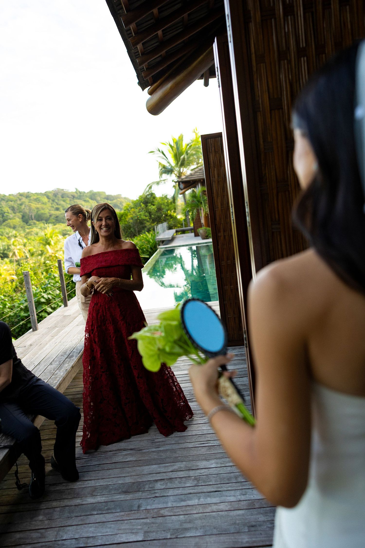 Bride in white dress holds bouquet while bridesmaid in red gown and groomsman stand on tropical villa deck.