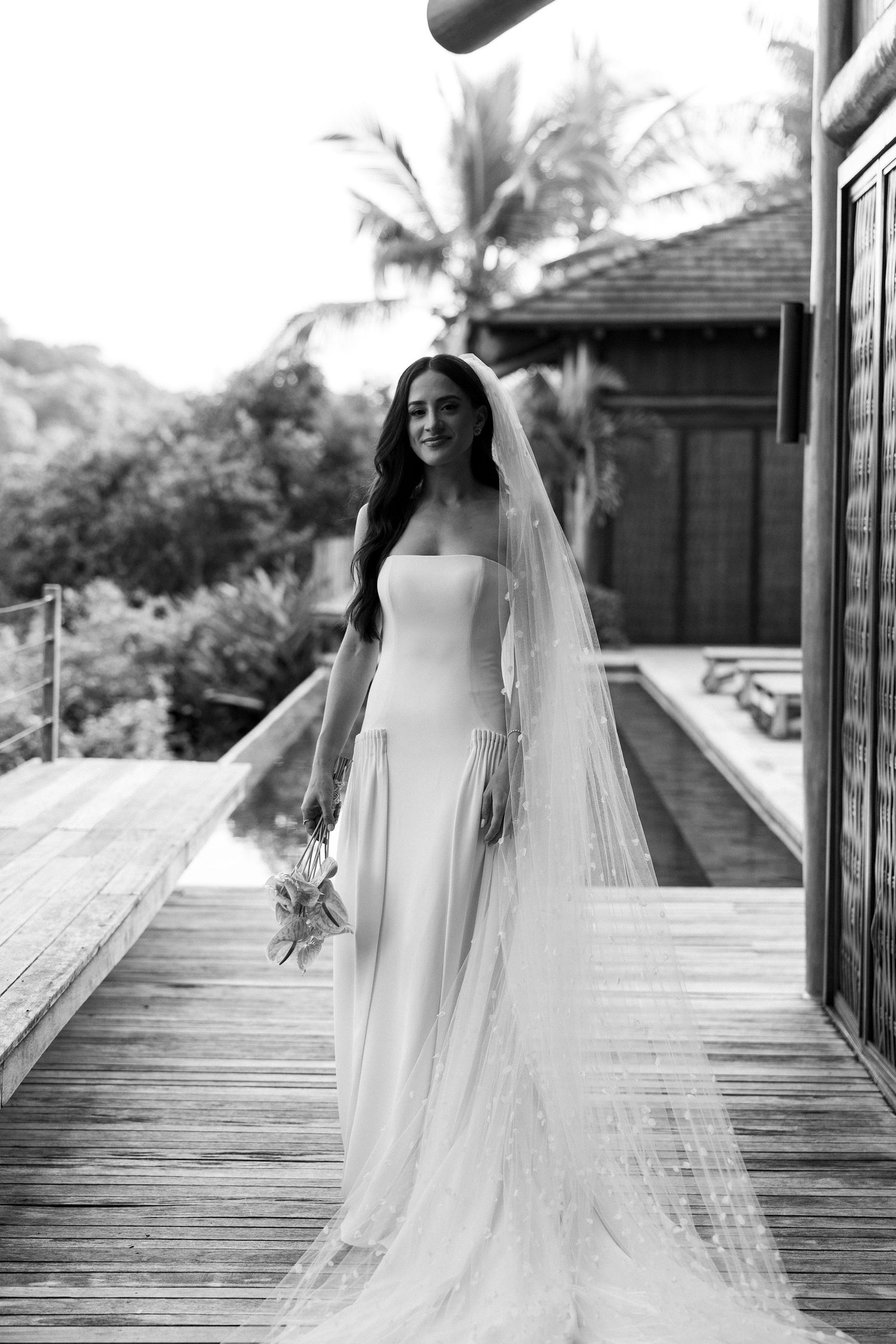 Black and white photo of a bride in a strapless gown and long veil on a wooden deck surrounded by tropical greenery.