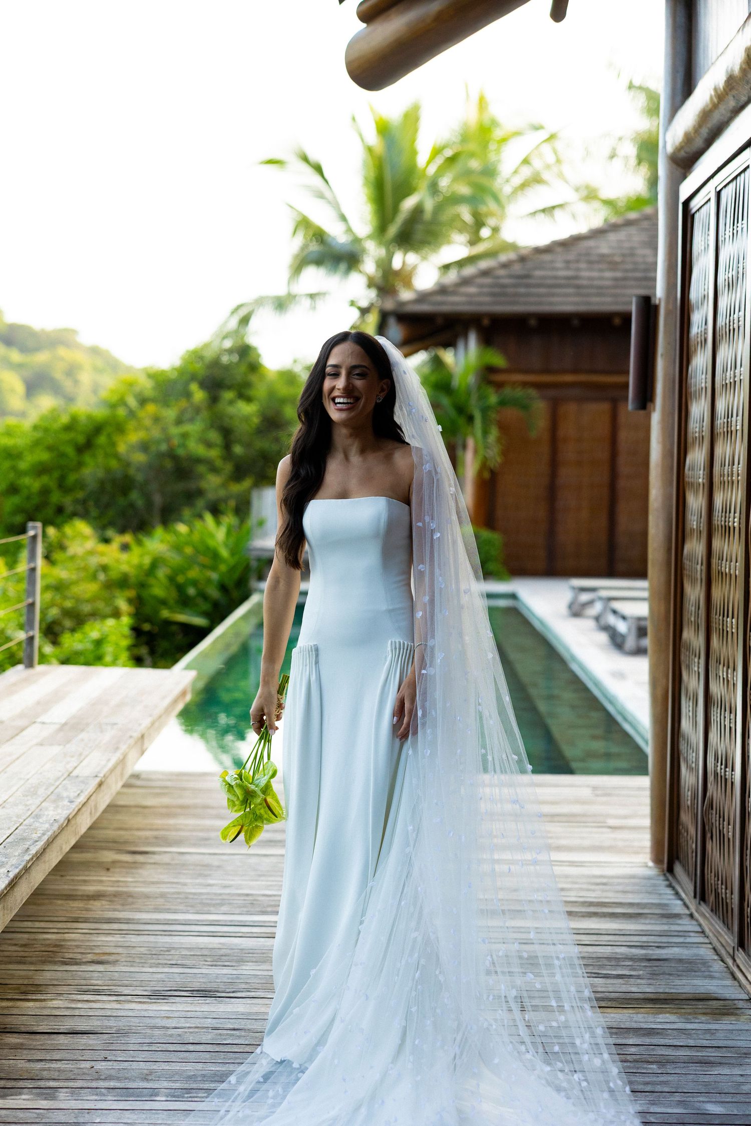 Laughing bride in elegant strapless white dress with flowing veil holds bouquet on wooden deck at tropical villa.
