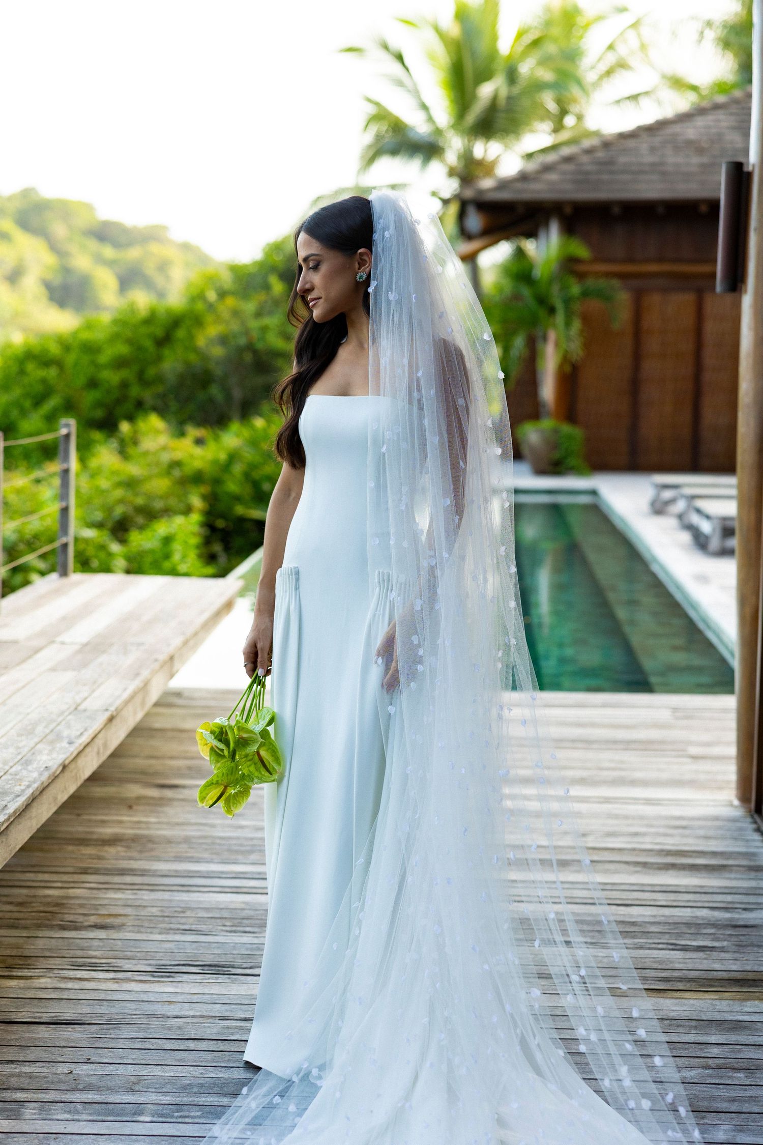 Bride in white gown and long veil holds green bouquet on wooden deck near pool with tropical greenery backdrop.