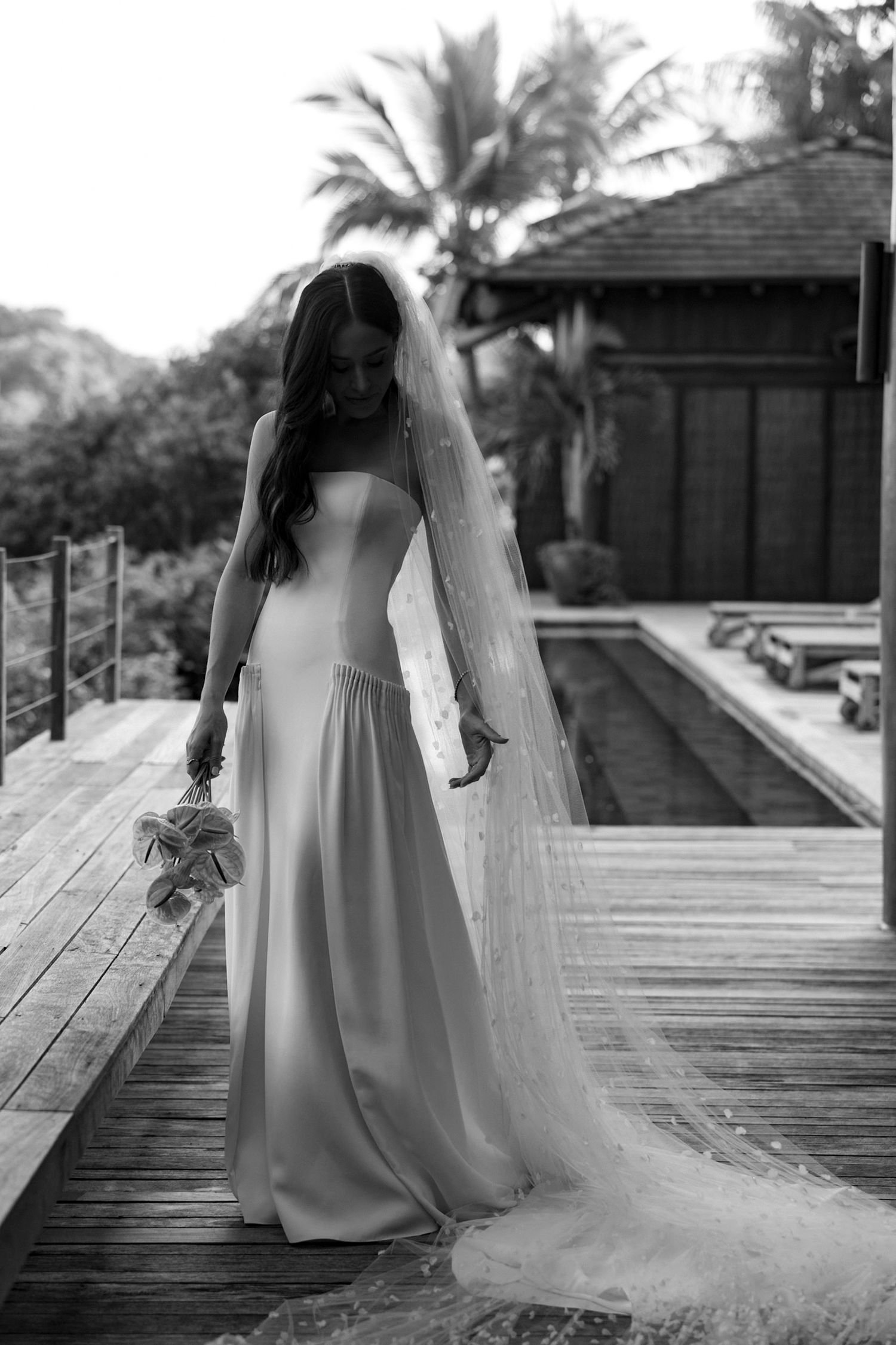 Bride in strapless white gown and long veil holds bouquet on wooden deck with pool and tropical foliage backdrop.