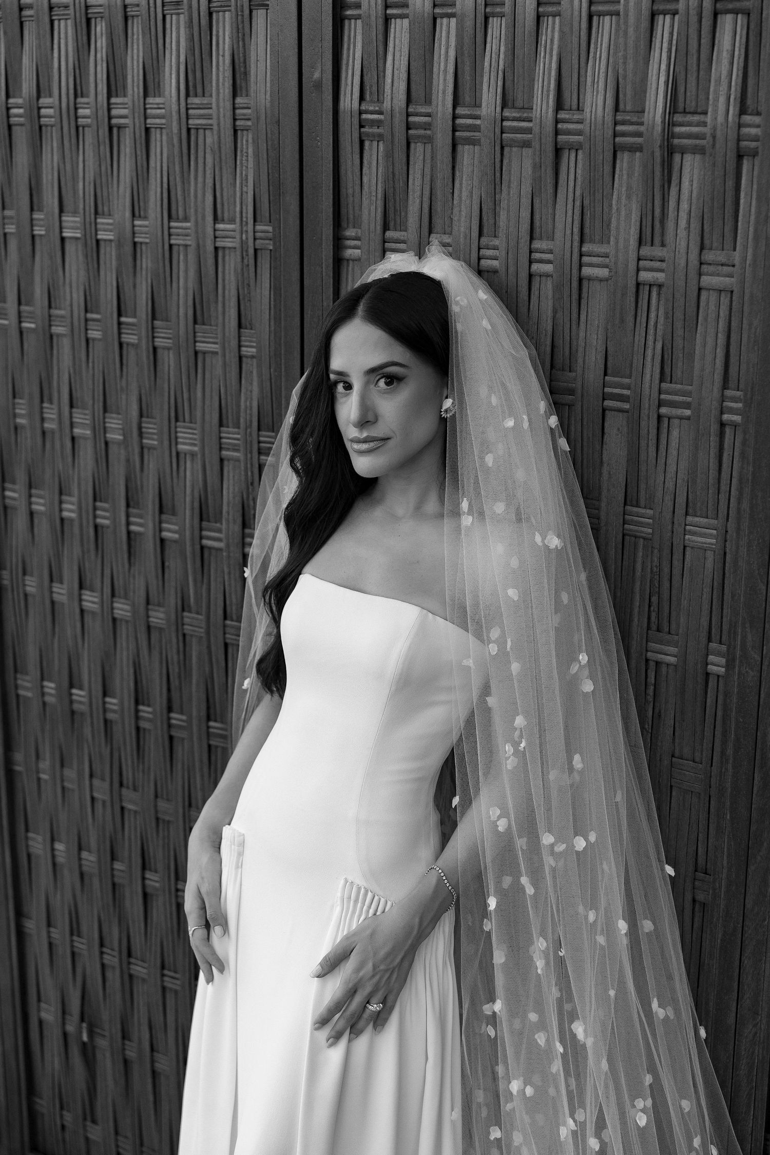 Black and white photo of a bride in a strapless gown and floral veil, posing elegantly against a woven bamboo wall.