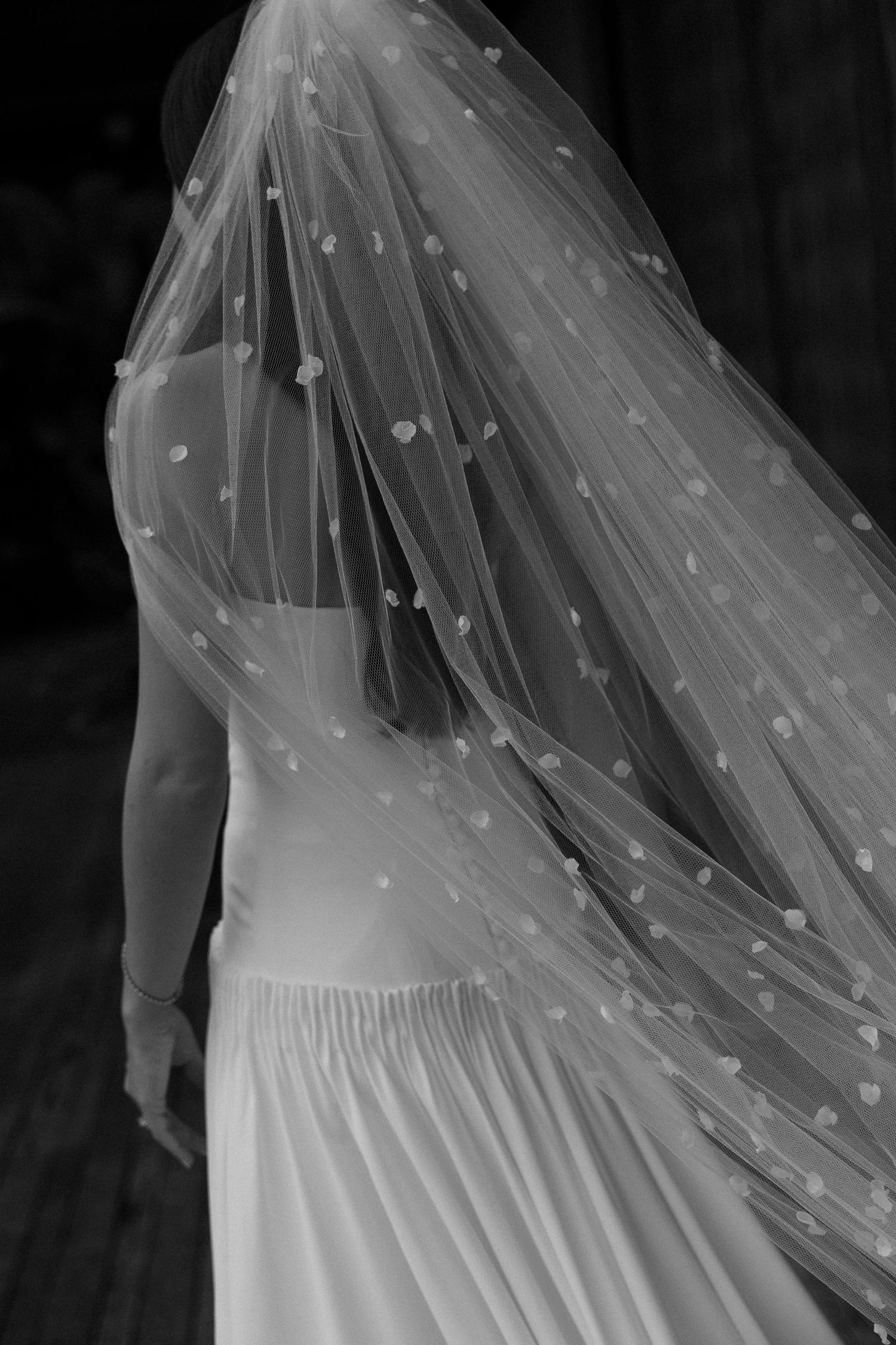 Black and white photo of a bride wearing a floral dotted veil and white gown, viewed from behind.