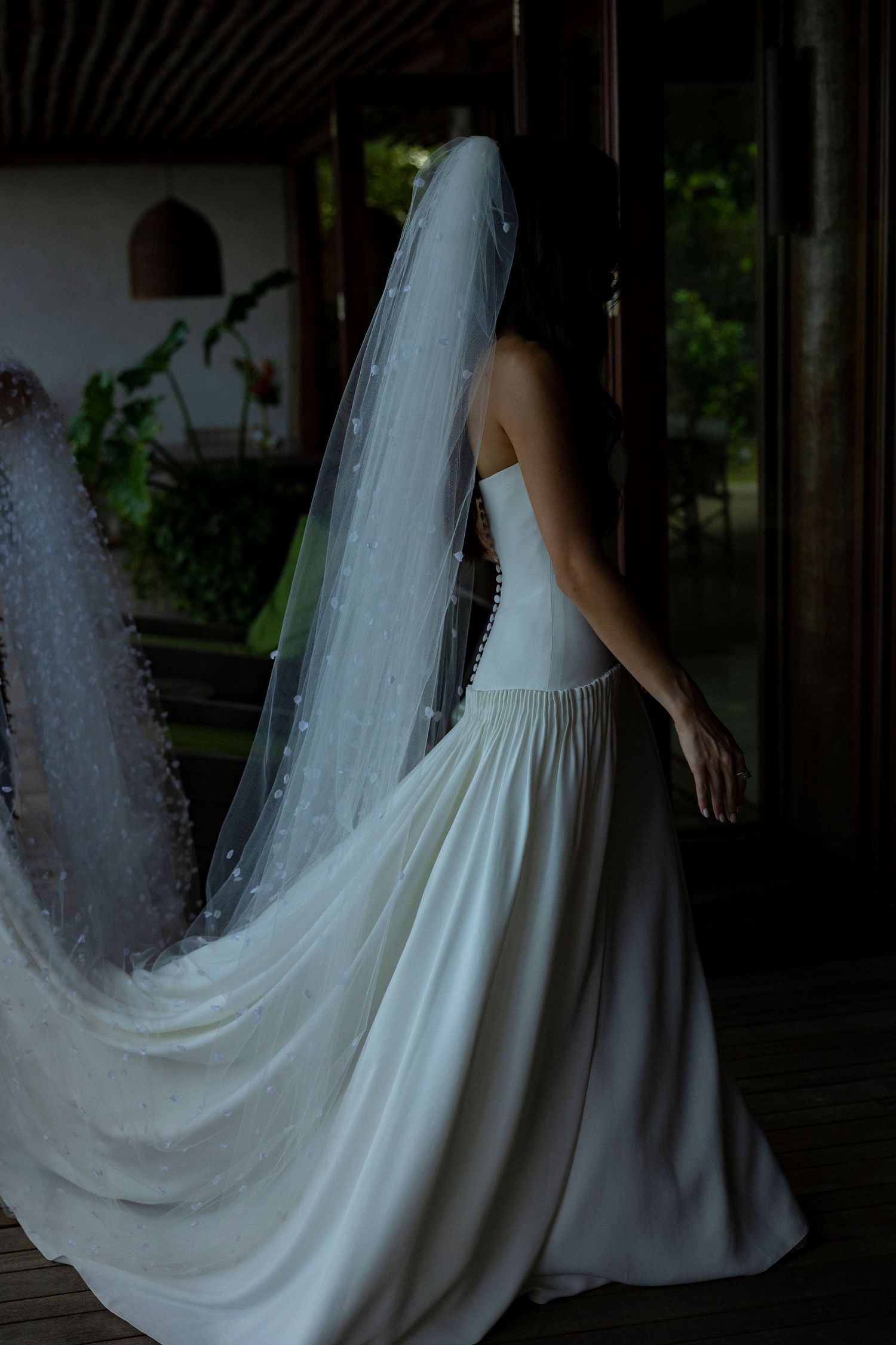 Bride in strapless white wedding gown with long flowing veil standing in doorway surrounded by lush greenery.