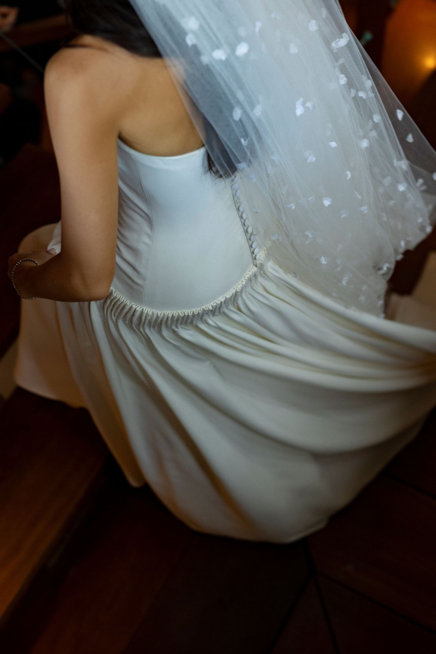 Bride in strapless white wedding dress with floral veil, photographed from behind in warm indoor lighting.