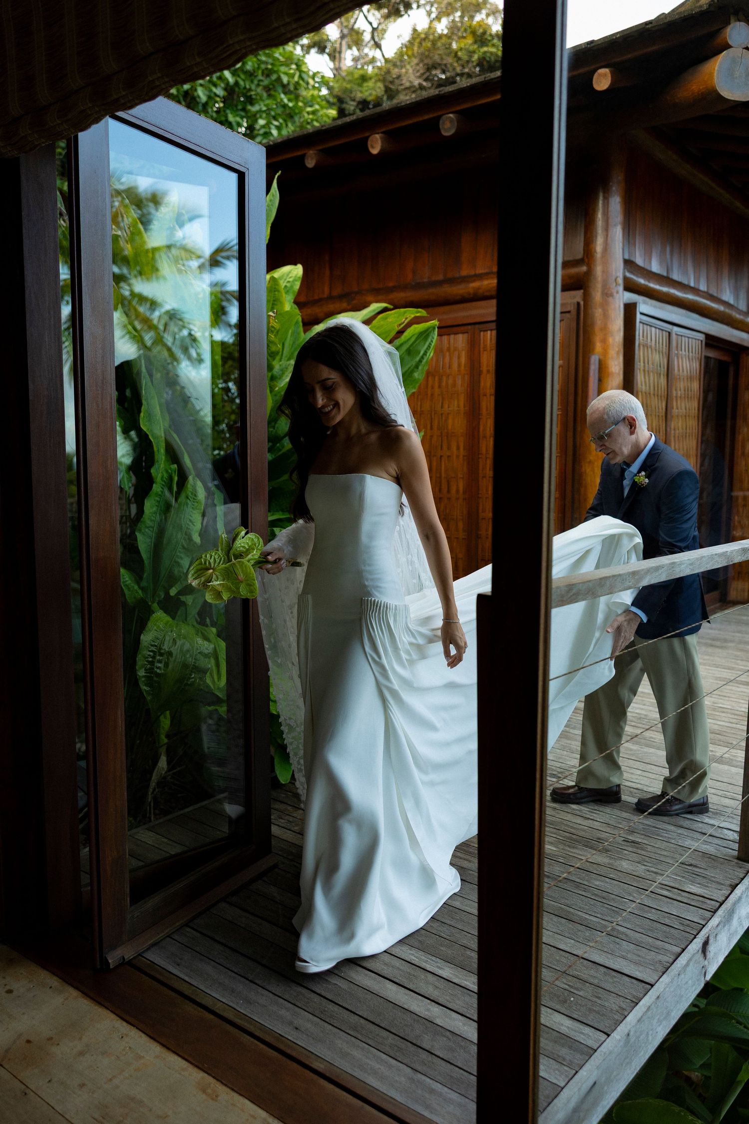 Bride in white strapless gown and veil walks through glass doors as man holds her train at tropical venue.
