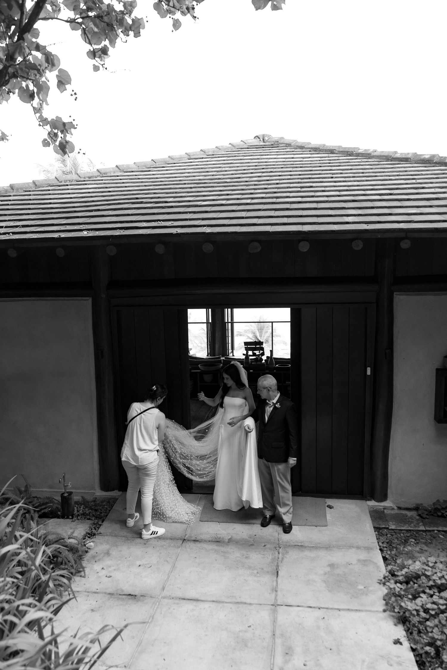 Black and white photo of a bride in a wedding gown being assisted with her train at a rustic venue entrance.