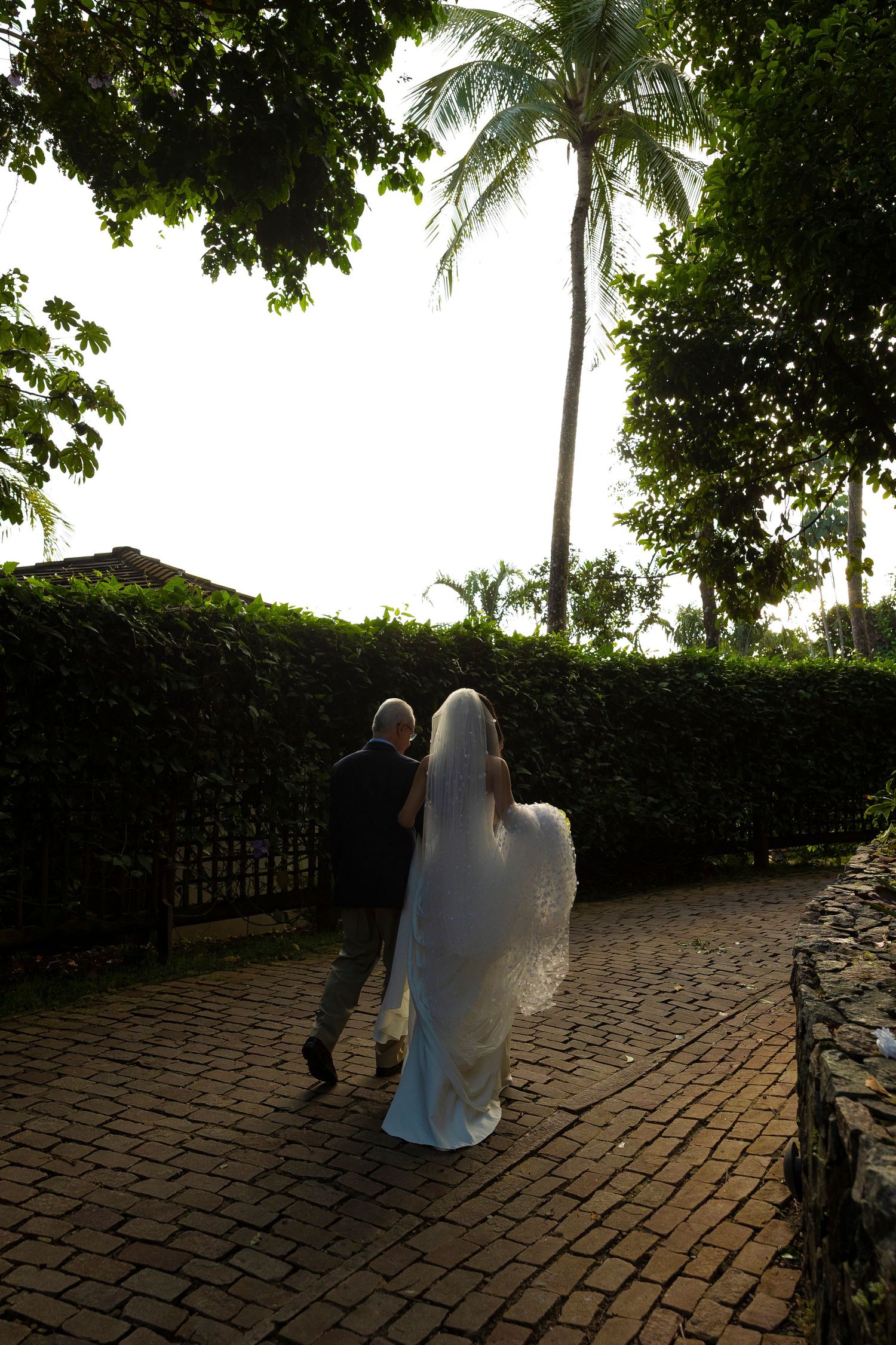 Bride and groom walking hand in hand along a brick path surrounded by lush tropical greenery and palm trees.