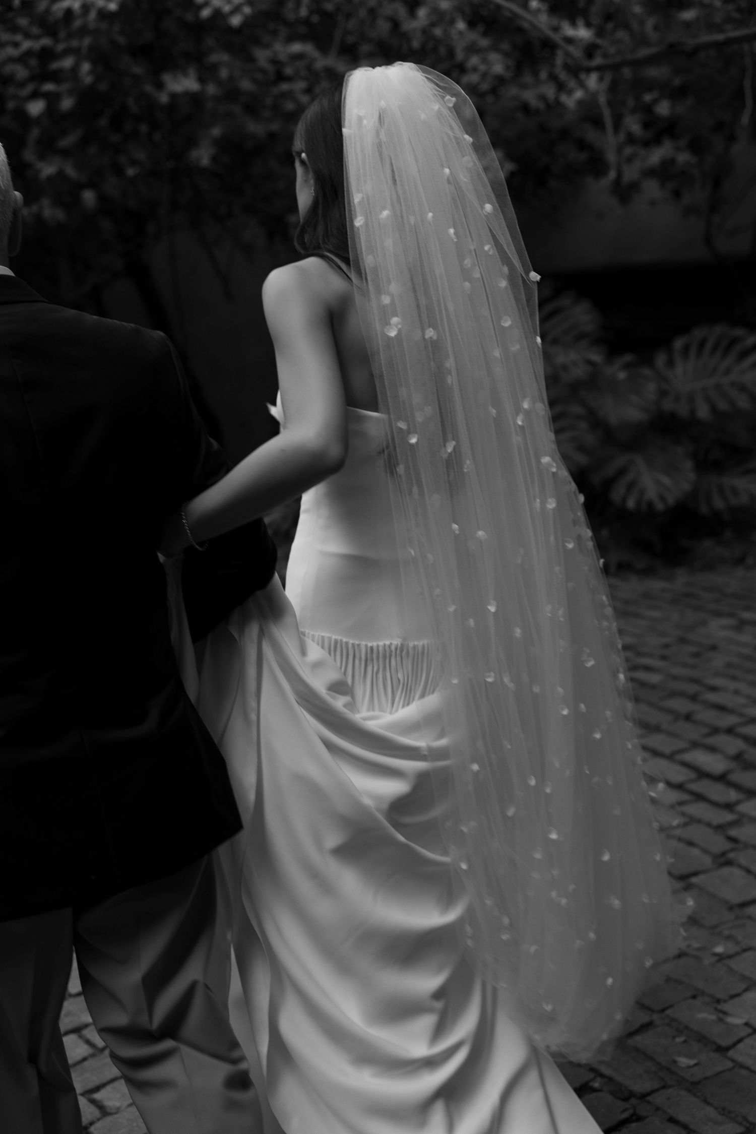 Black and white photo of a bride in a white gown and floral veil walking with a groom on cobblestone.