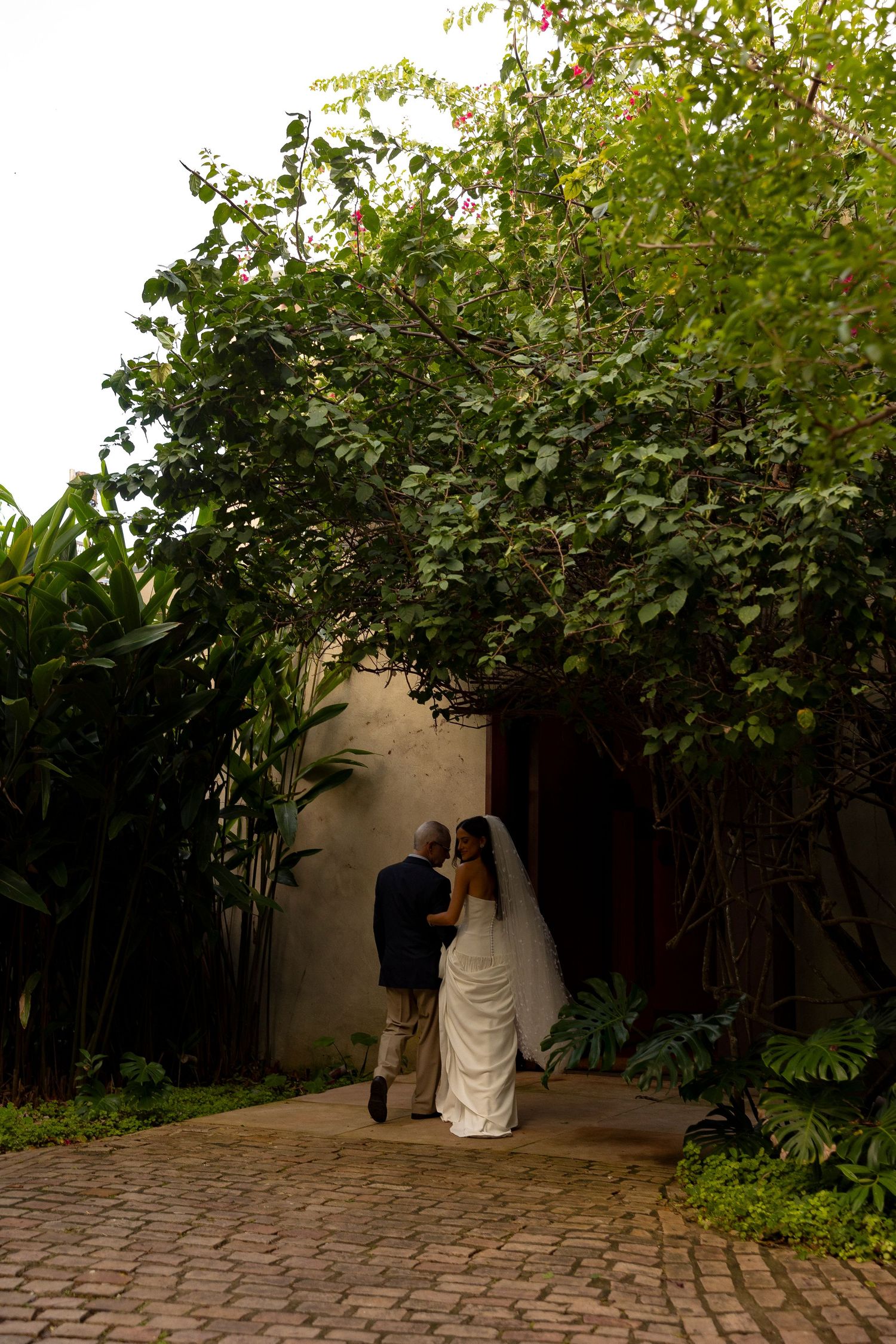 Bride and groom embrace under a lush green archway on a cobblestone path at an outdoor garden wedding venue.