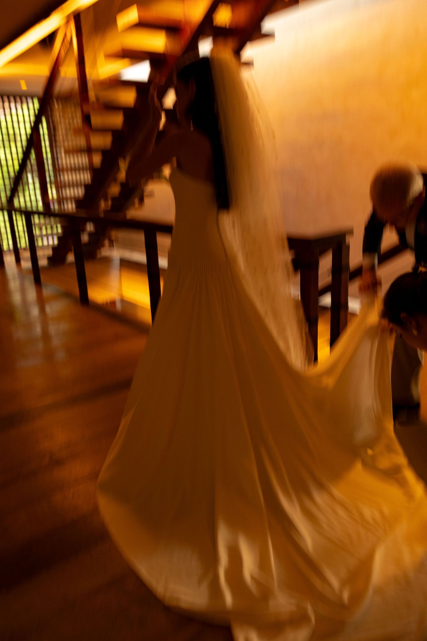 Bride in flowing white wedding gown and veil captured in motion near staircase in warm golden light.