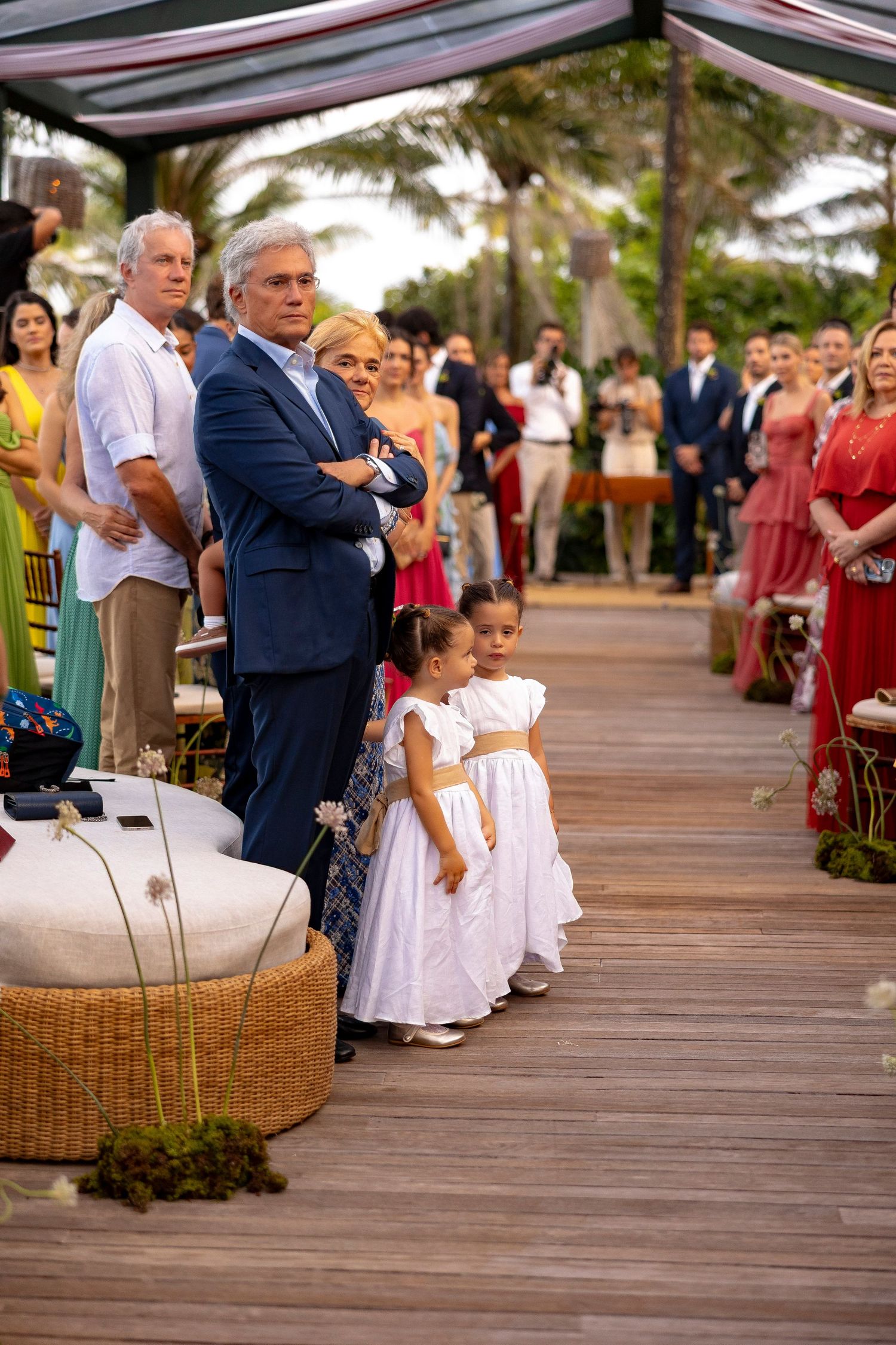Two flower girls in white dresses stand at an outdoor wedding ceremony on a wooden deck surrounded by guests.