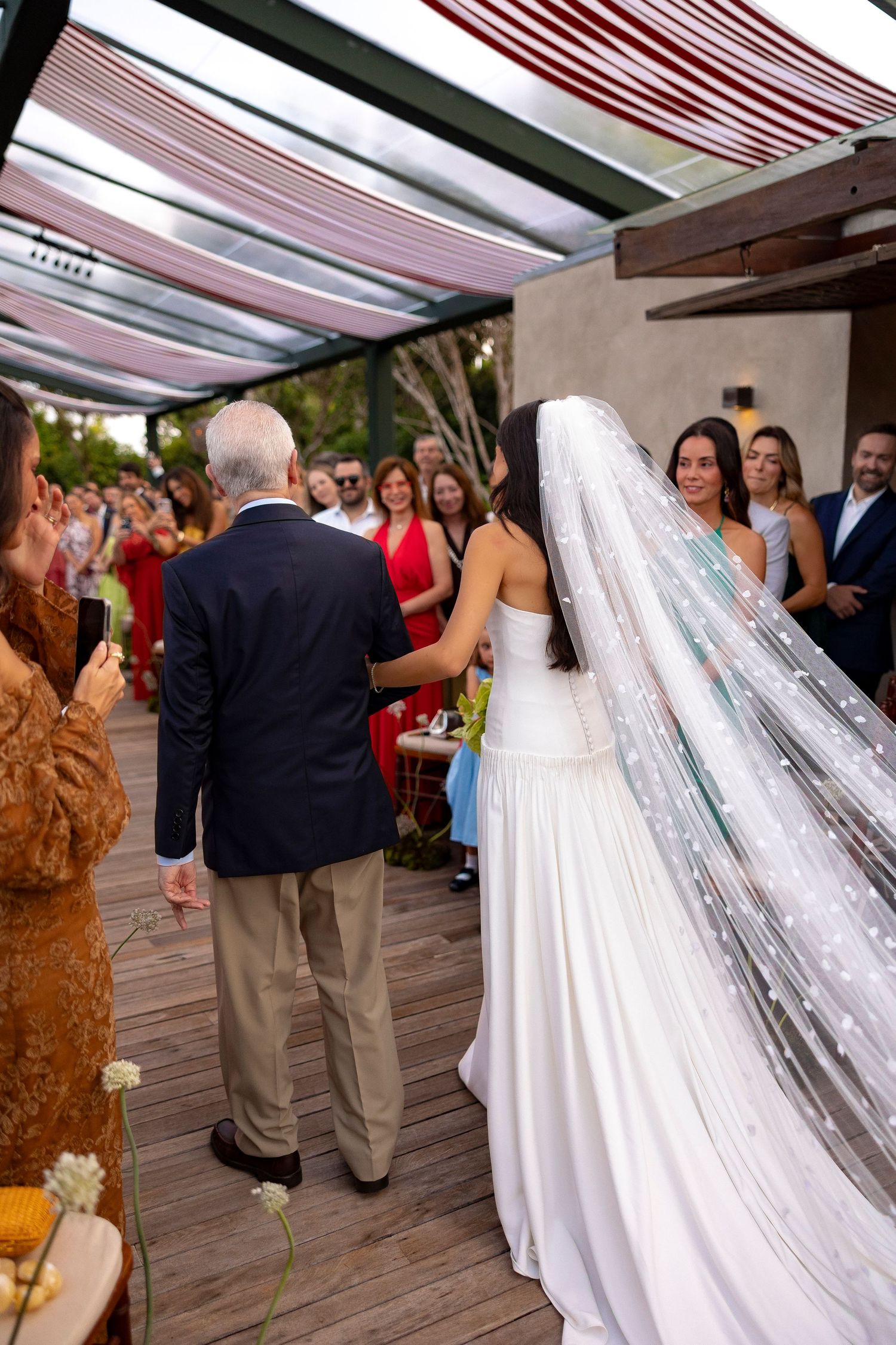 Bride in white gown and veil walks with elderly man at outdoor wedding ceremony with guests watching.