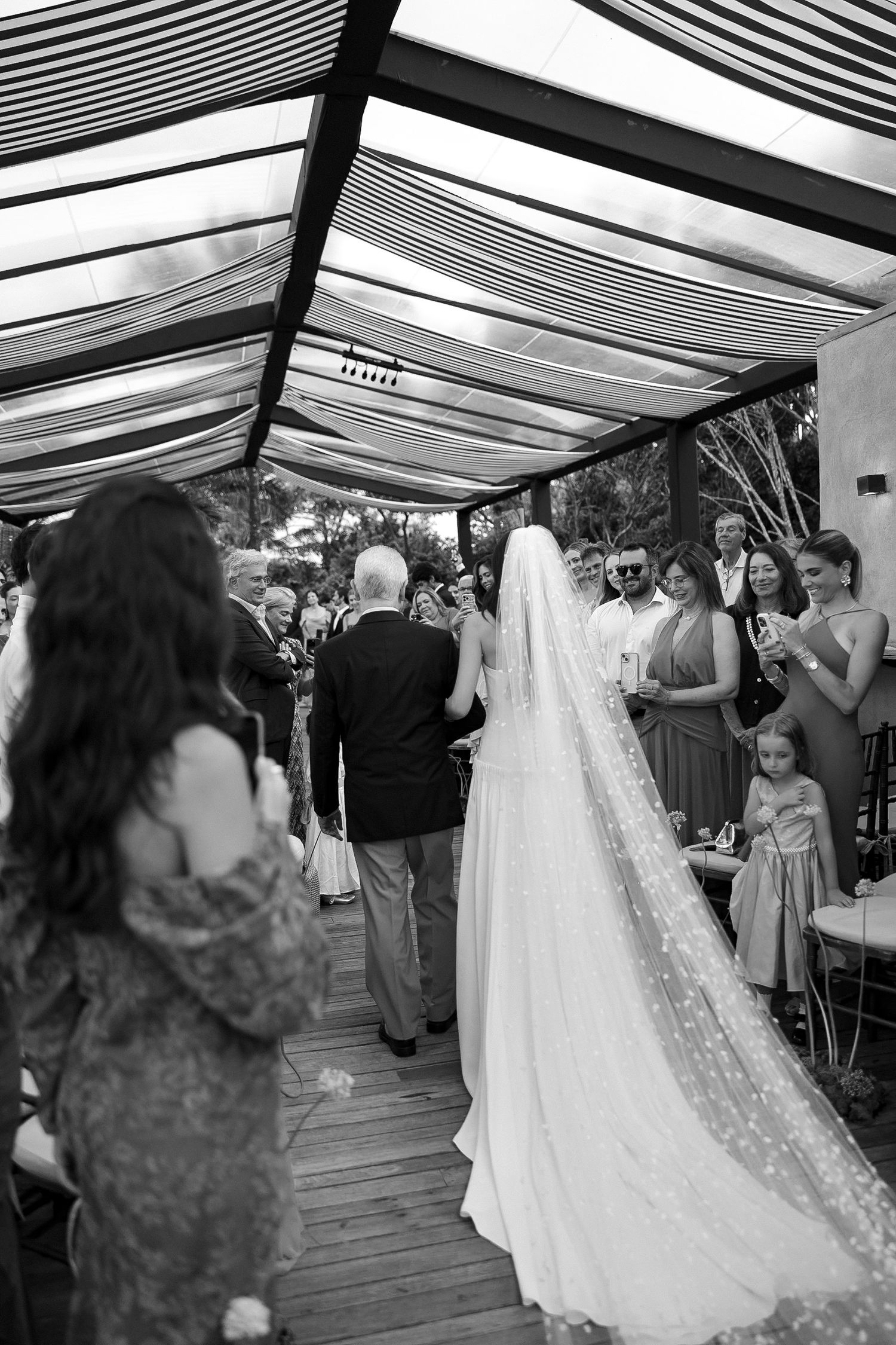 Black and white photo of a bride in a long veil walking down the aisle at an outdoor wedding ceremony.