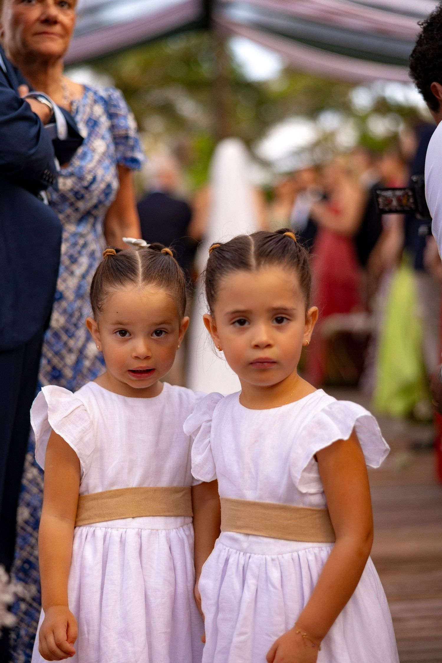 Two young twin girls in matching white dresses with gold sashes stand together at a formal outdoor event.