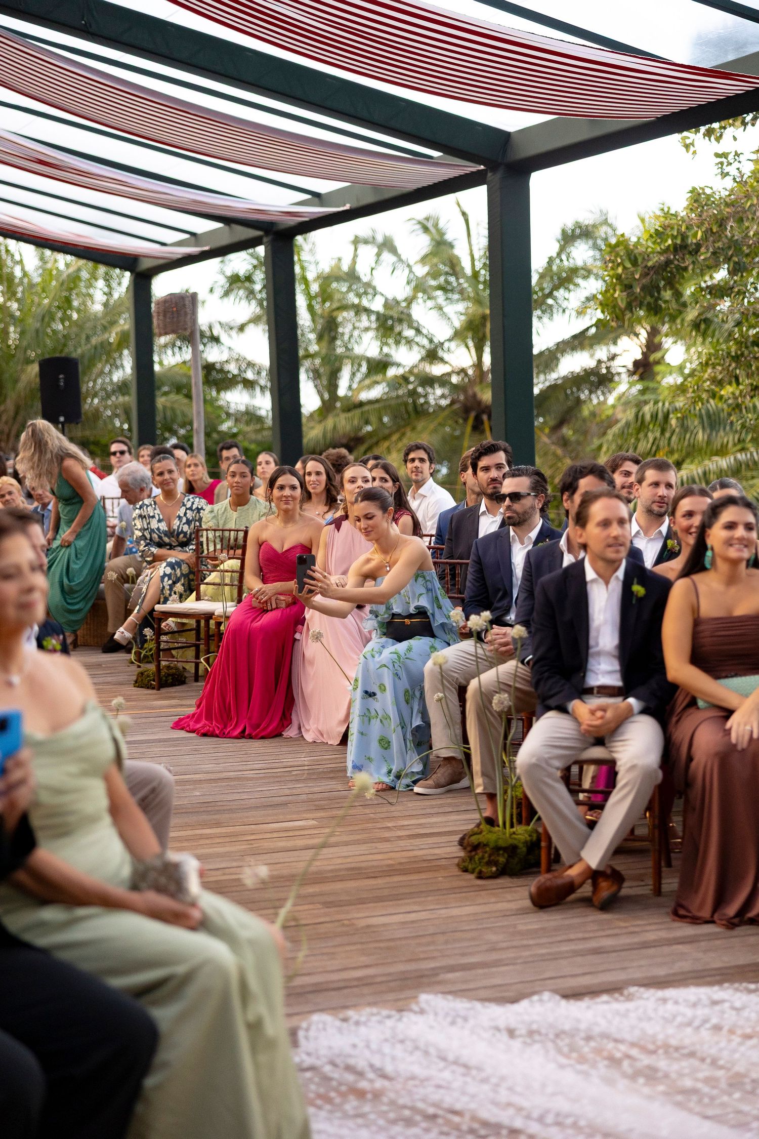 Guests seated outdoors at an elegant wedding ceremony on a wooden deck surrounded by tropical greenery.