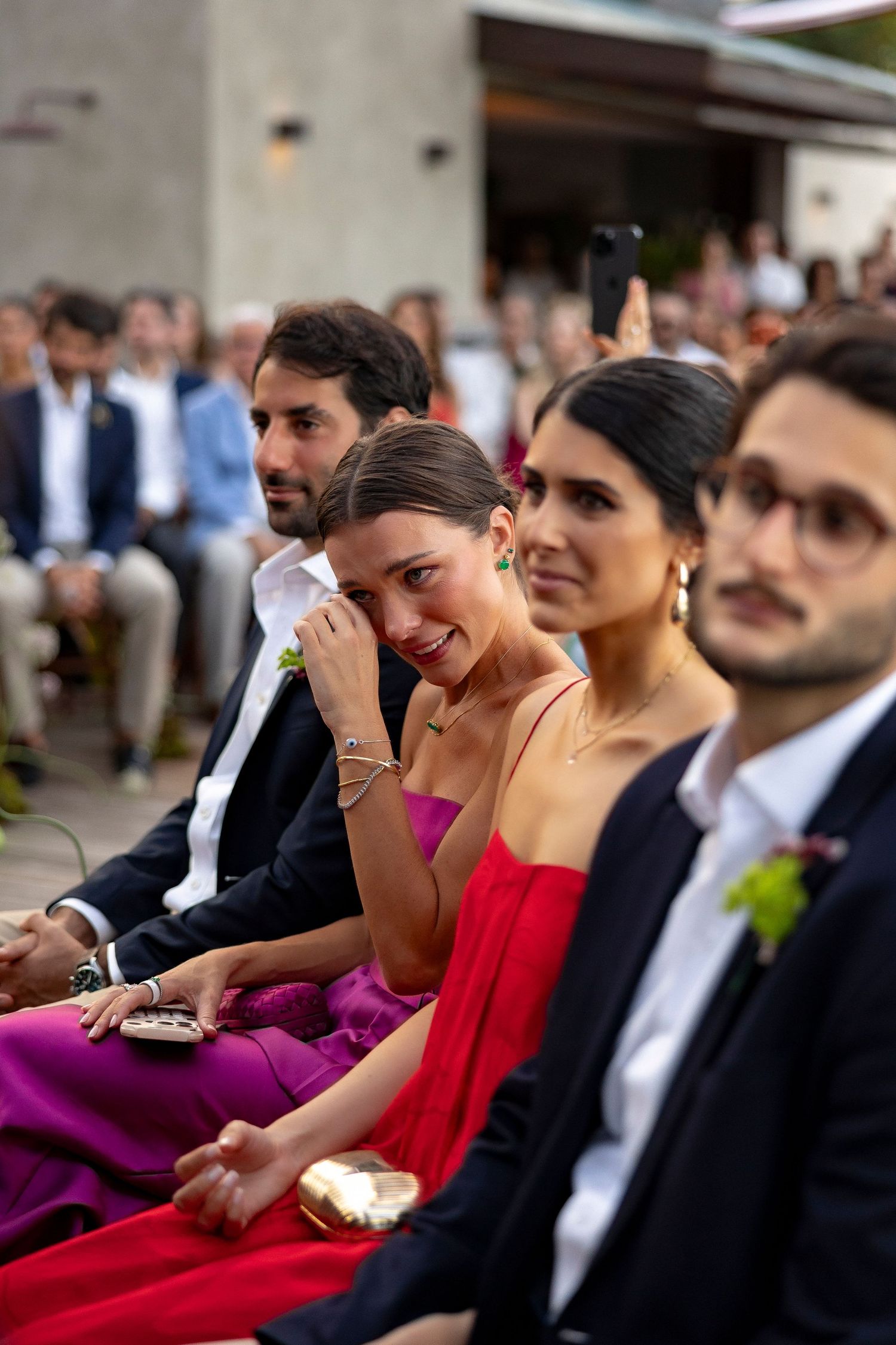 Emotional wedding guest in purple dress wiping tears during outdoor ceremony, seated among elegantly dressed attendees.