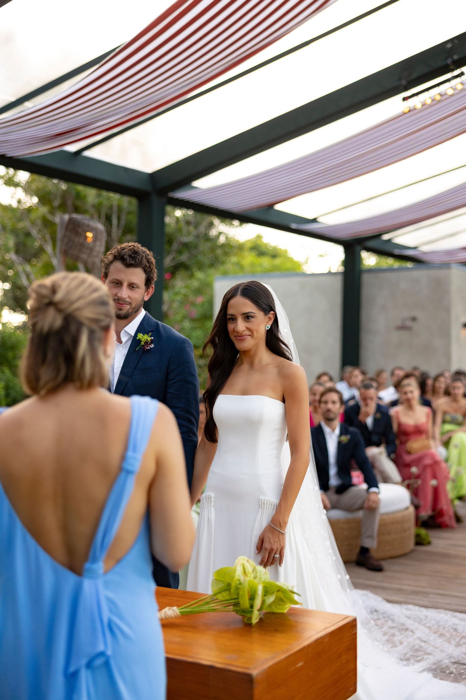 Bride in white strapless gown and groom in navy suit exchange vows at an outdoor wedding ceremony under a modern pergola.