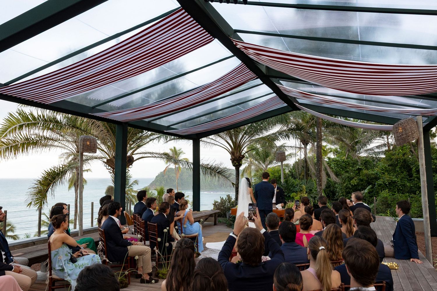 Outdoor wedding ceremony under a striped canopy with ocean views and tropical palm trees in the background.